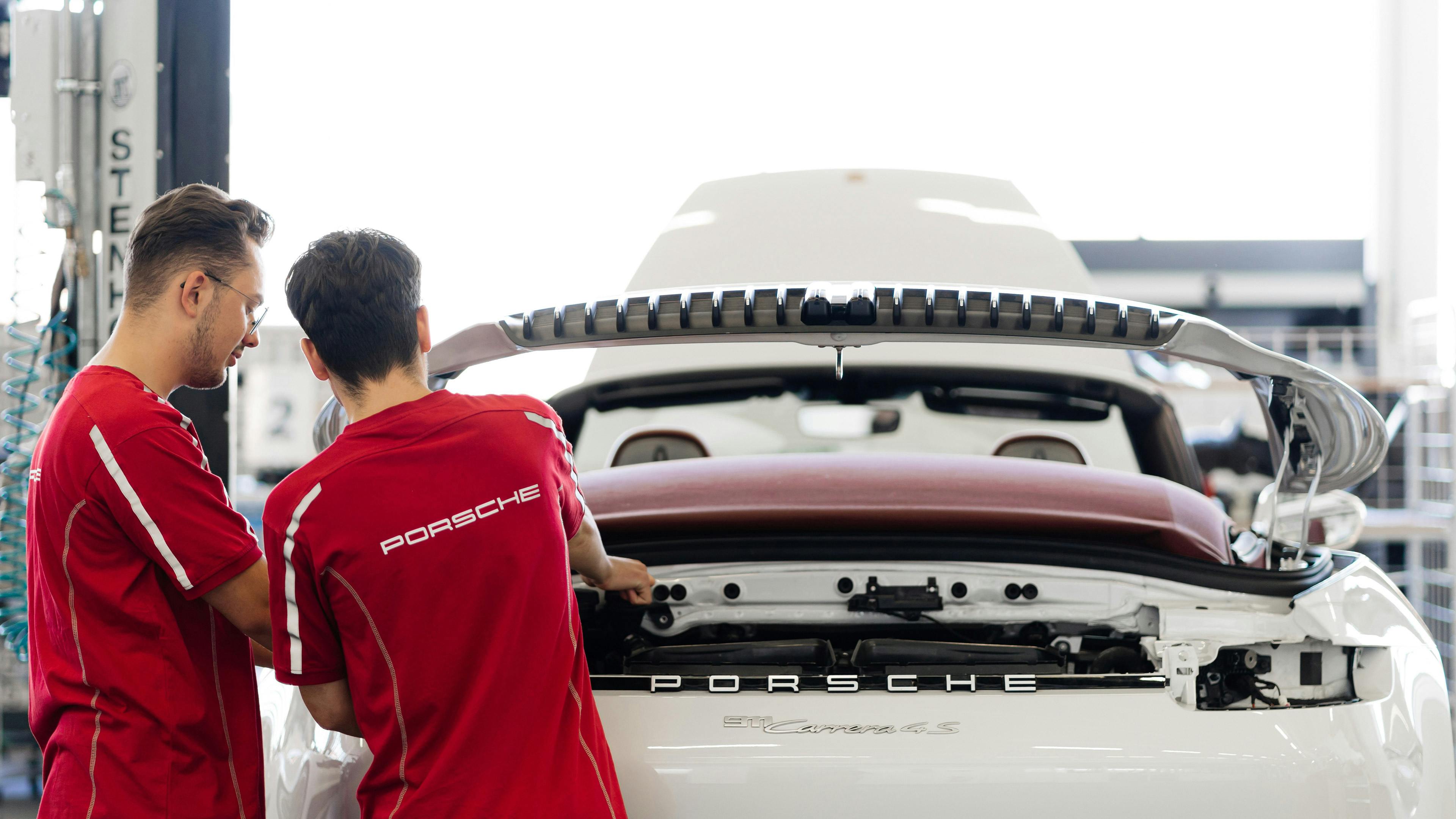 Zwei Porsche Auszubildende arbeiten gemeinsam am geöffneten Heck eines weißen Porsche‑Fahrzeugs in einer Werkstattumgebung.