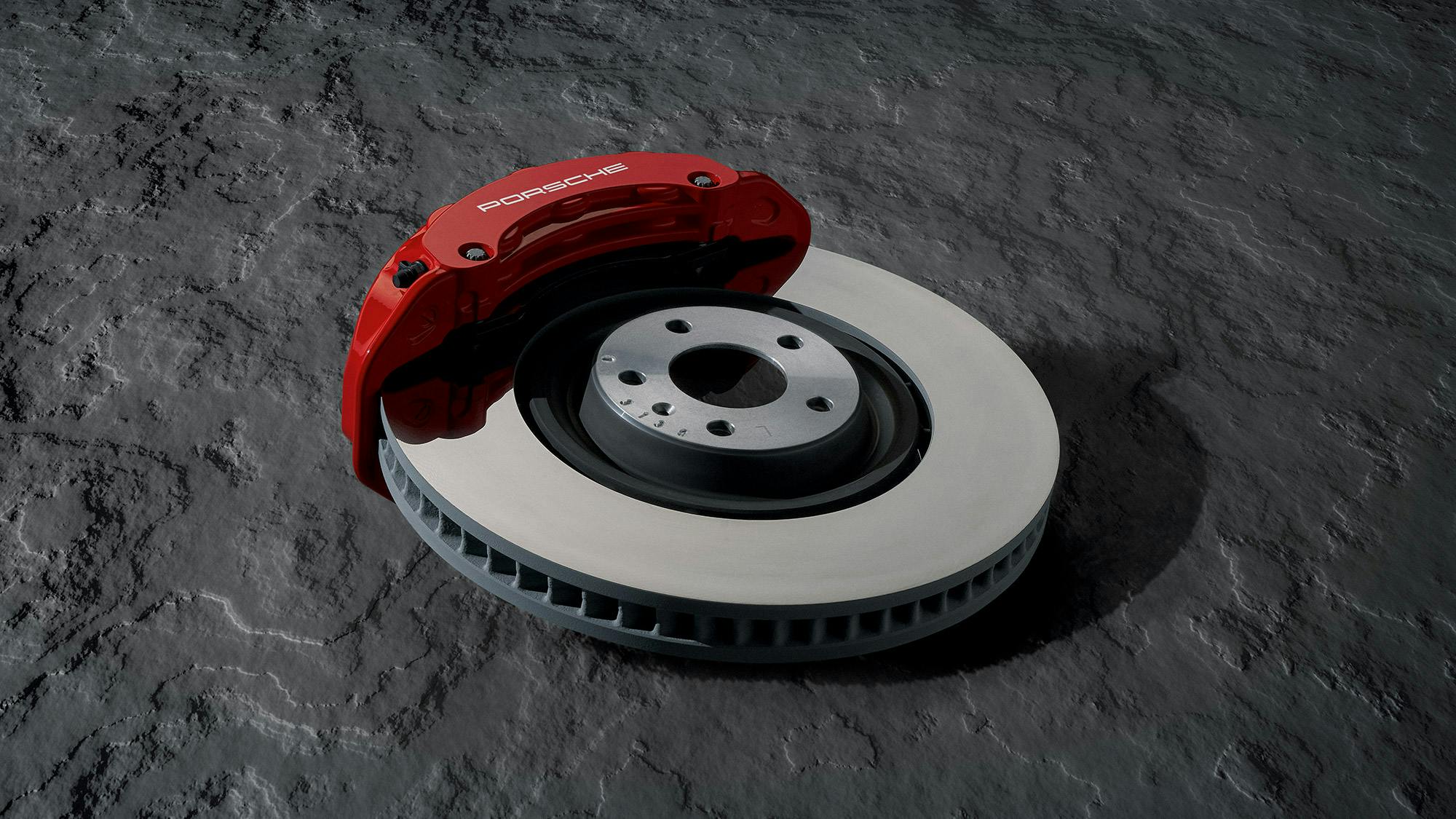 A red Porsche brake calliper and a perforated brake disc lie on a textured concrete slab.