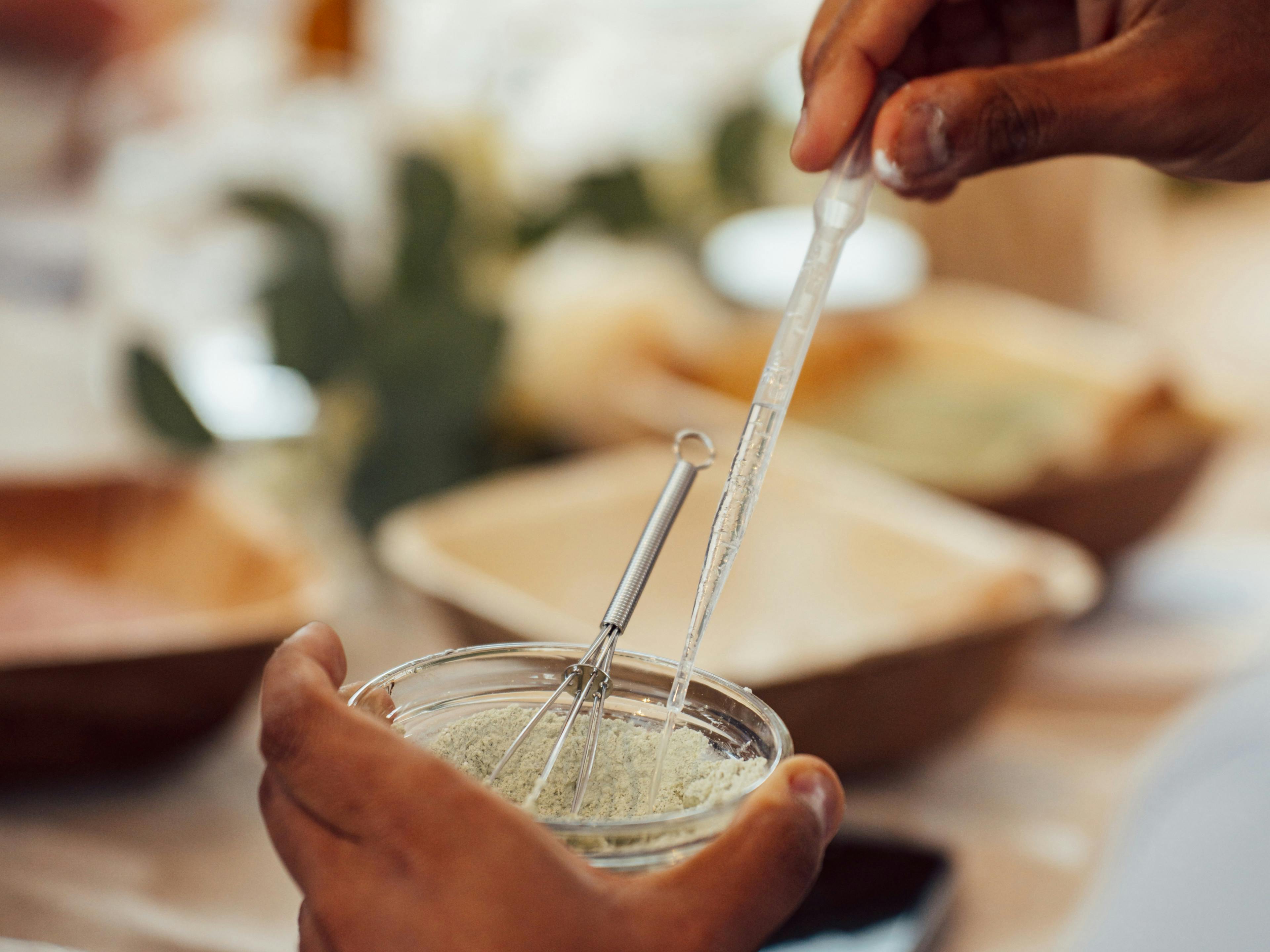 A person’s hands are seen using a pipette and a small whisk to mix a light-coloured powder in a glass bowl.​