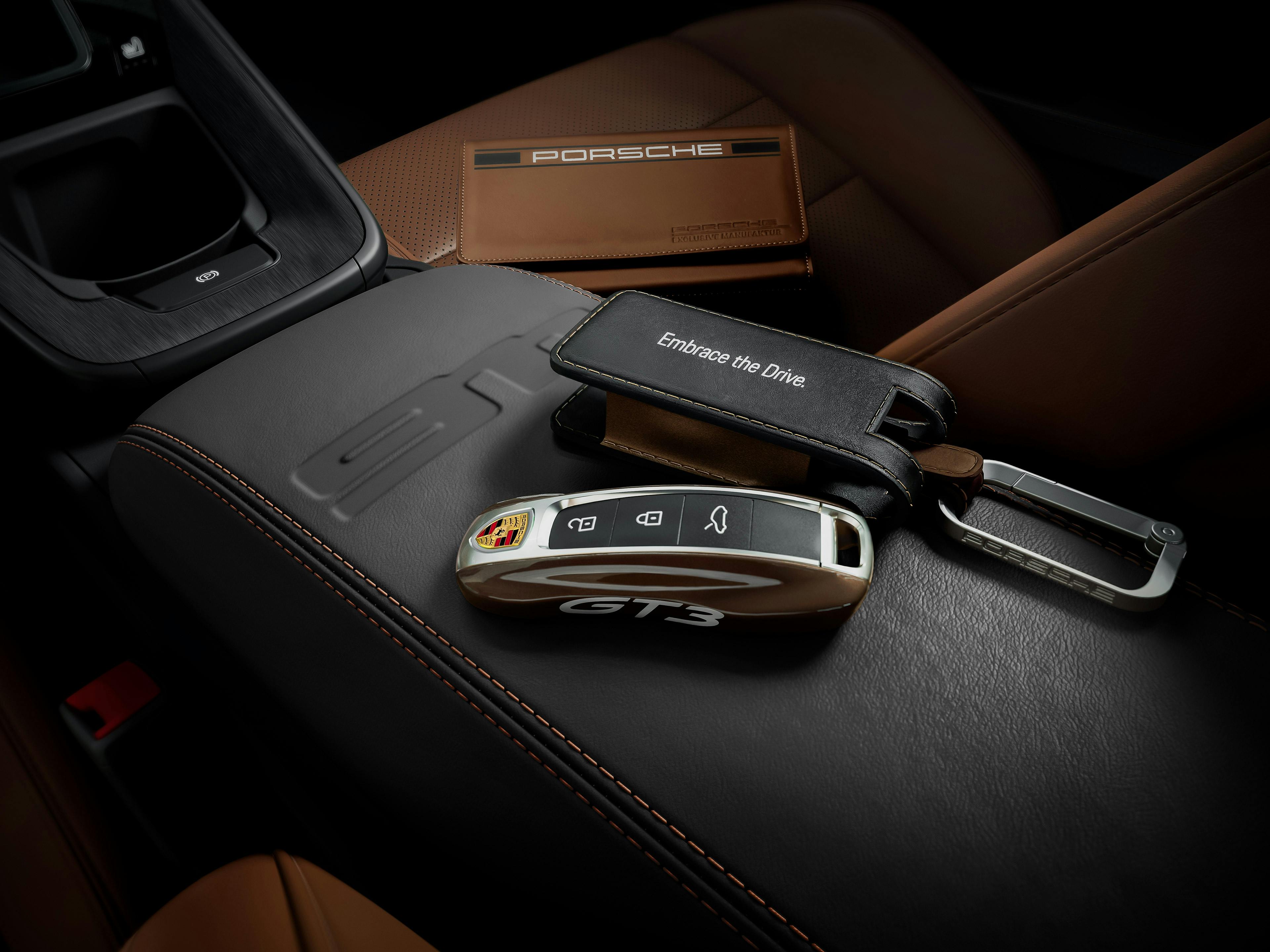 Ein Porsche GT3-Autoschlüssel mit schwarzem Schlüsselanhänger, auf dem „Embrace the Drive.“ steht, liegt auf einer schwarzen Leder-Mittelarmlehne mit orangefarbenen Ziernähten. Im Hintergrund sind braune Ledersitze und eine Porsche-Ledermappe sichtbar.