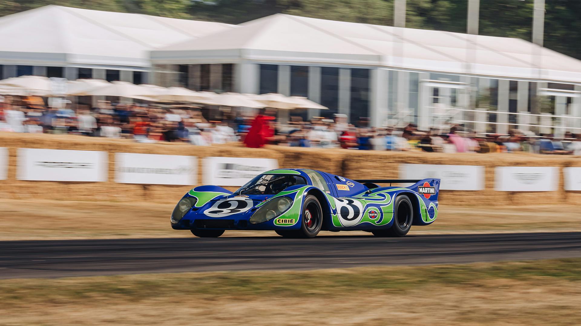 Two liveried Porsche race cars on track in front of the grandstand.