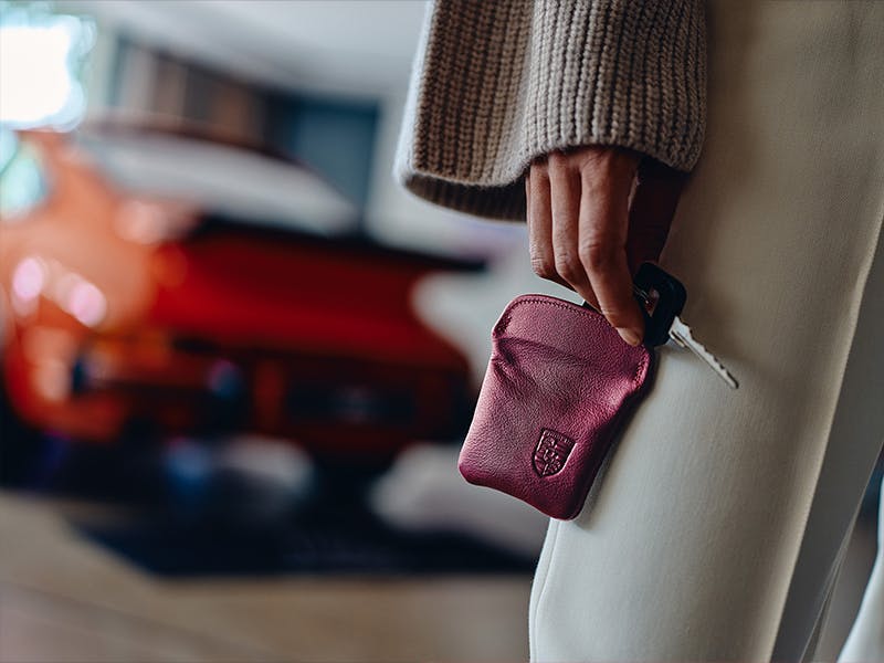 La femme tient un étui en cuir marron dans la main. Une Porsche se trouve à l'arrière-plan.