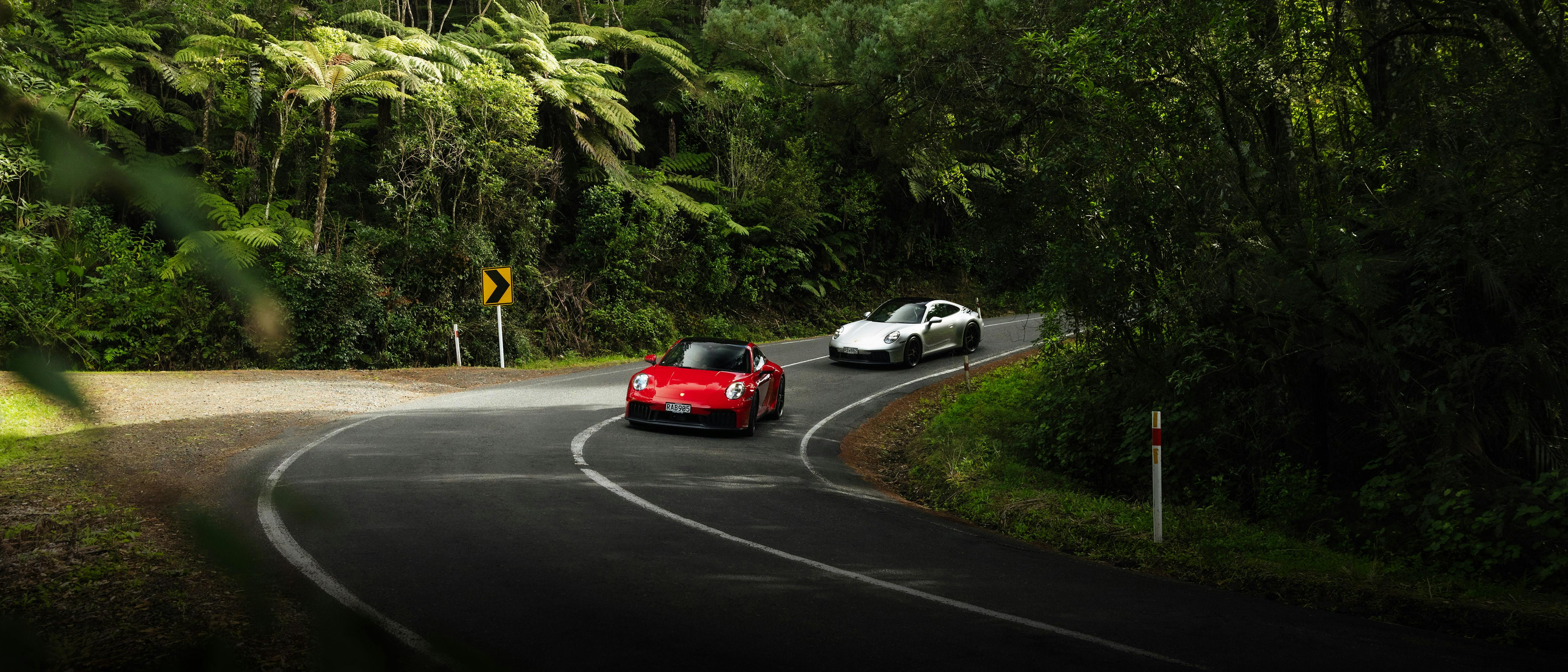 Two Porsches, one red and one silver, drive around a sharp bend on a forest road, surrounded by dense green vegetation.
