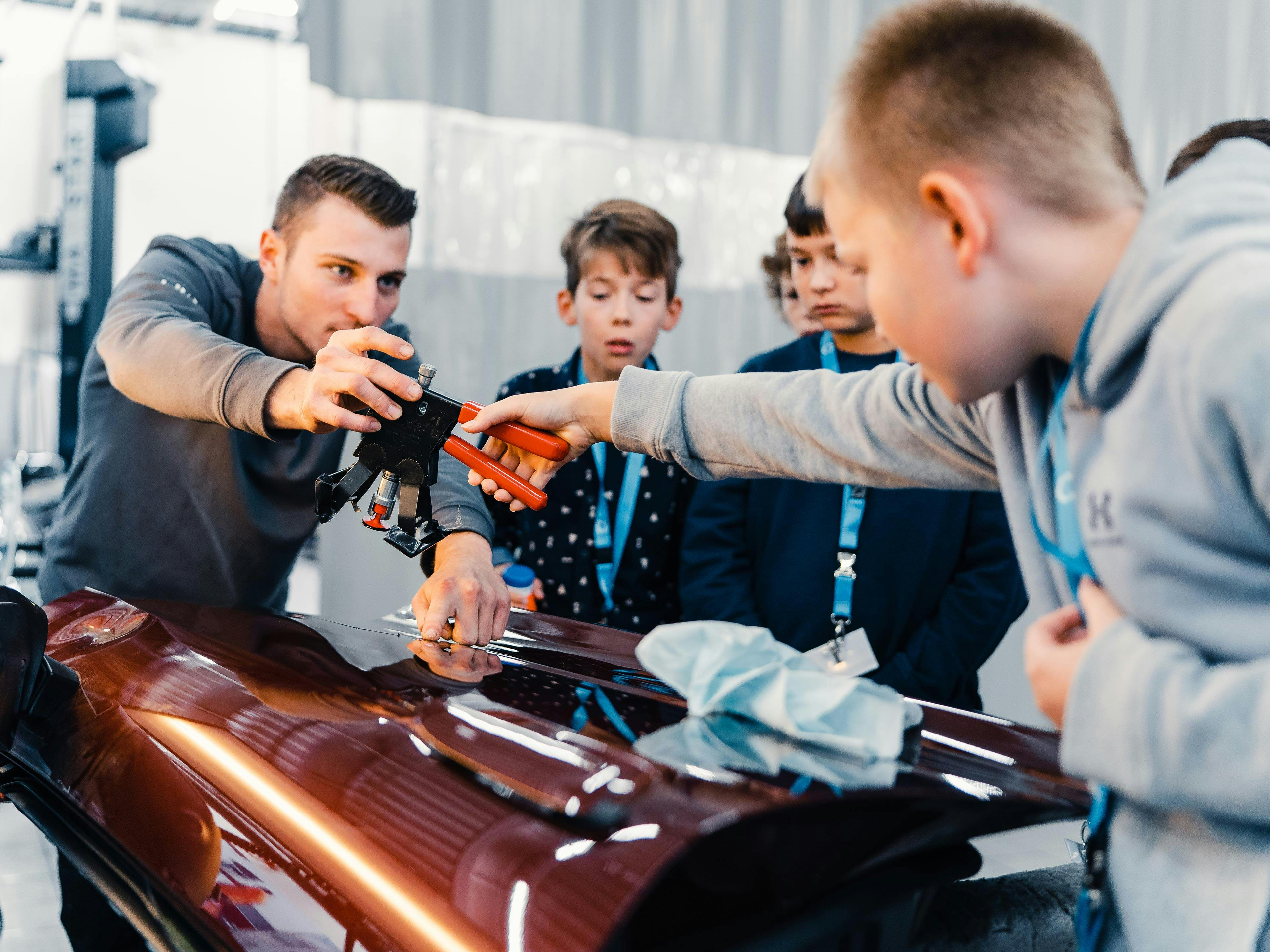 Ein Porsche Mitarbeiter zeigt einer Gruppe von Kindern, wie man ein Ausbeulgerät benutzt.