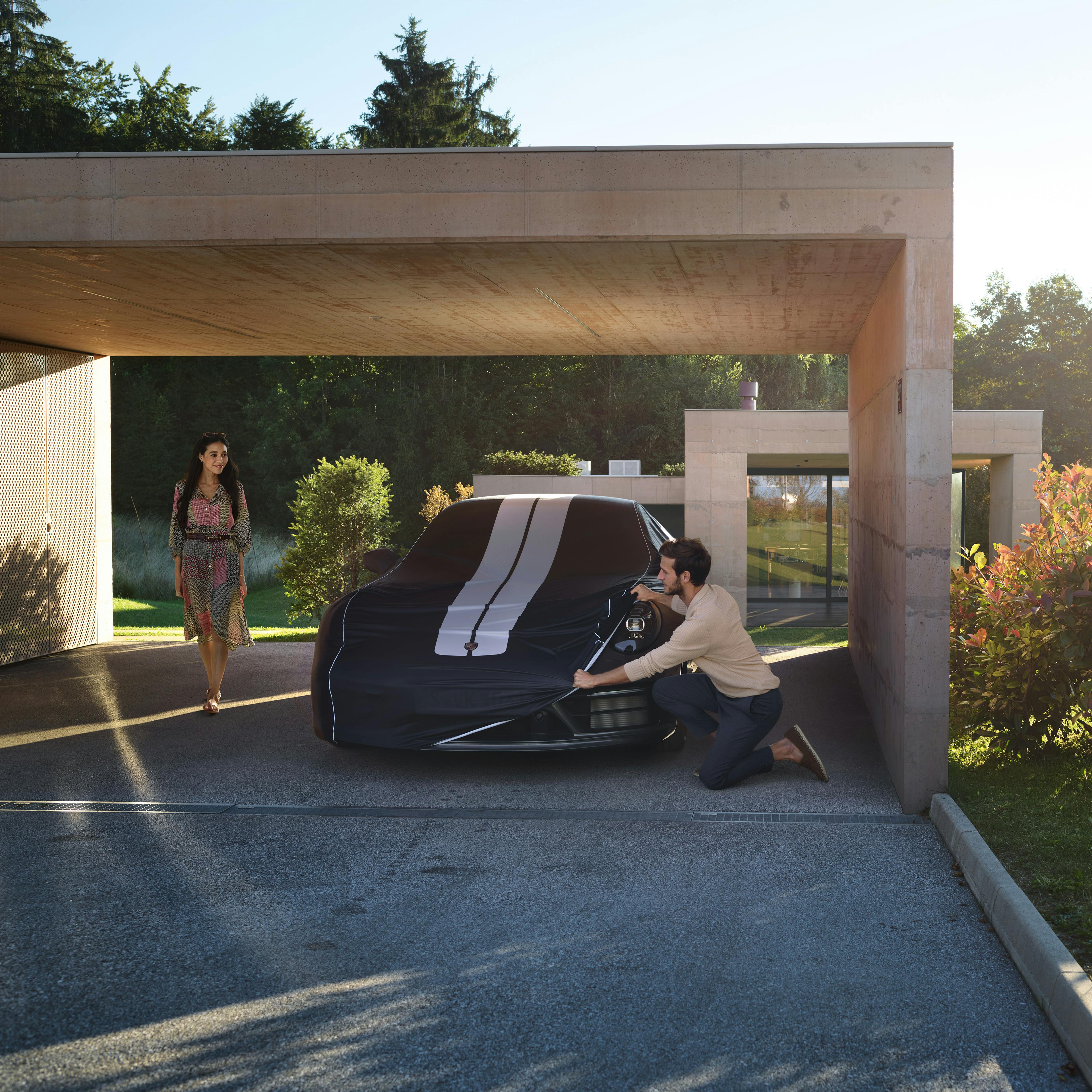 A Porsche 911 is being covered with a striped Indoor Car Cover Plus in a garage; there are two people in the garage.