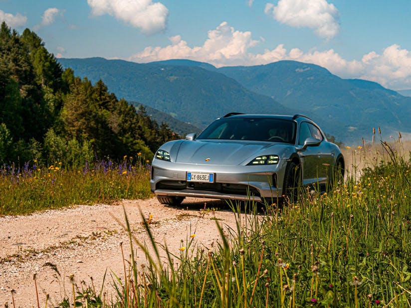 Silver Porsche Taycan Cross Turismo on gravel mountain road surrounded by wildflowers and meadow grass with forested mountains in background under blue sky