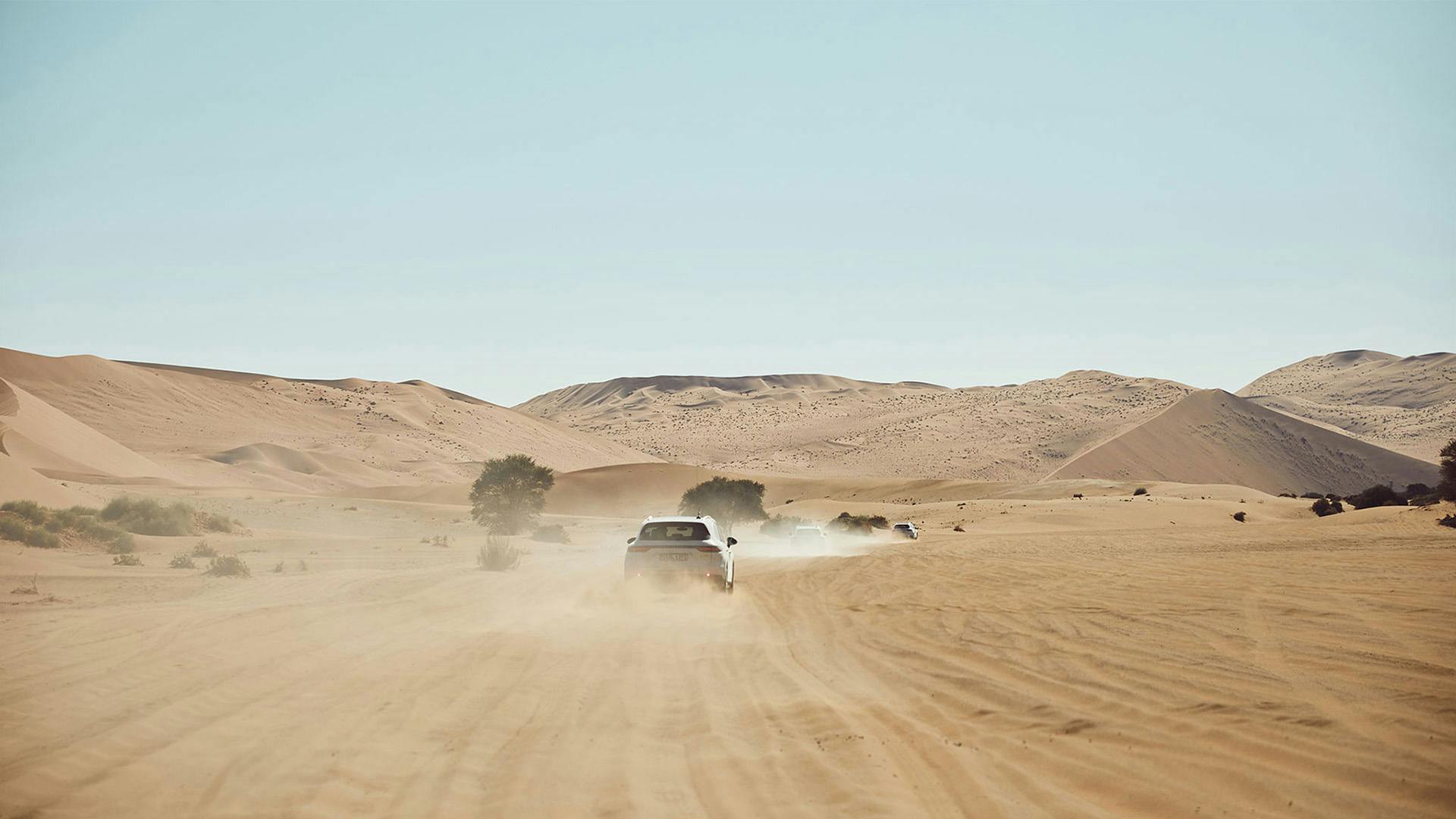 A Porsche kicks up sand and dust as it accelerates between dunes and hills in the middle of the desert landscape.