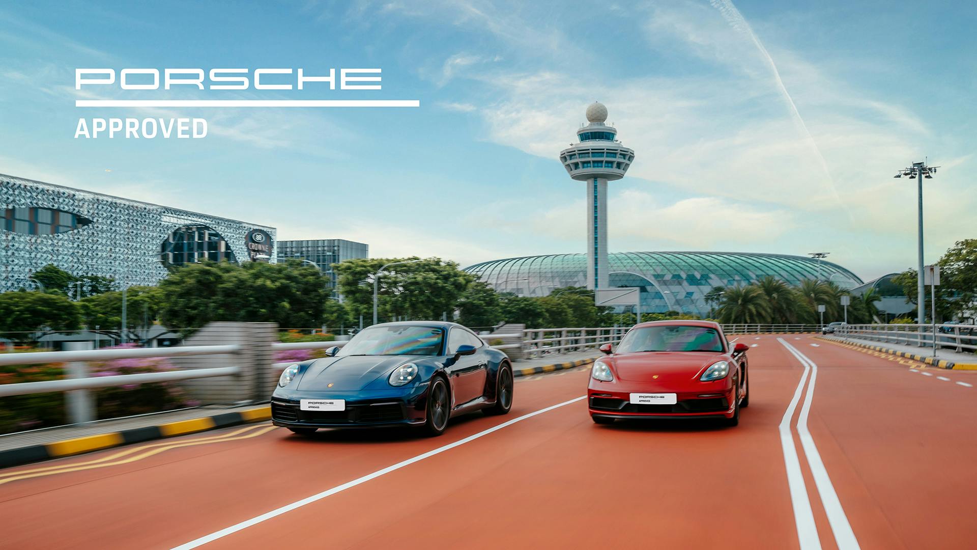 Two Porsche cars, blue and red, driving side by side past modern architecture and tower, with “Porsche Approved” text.