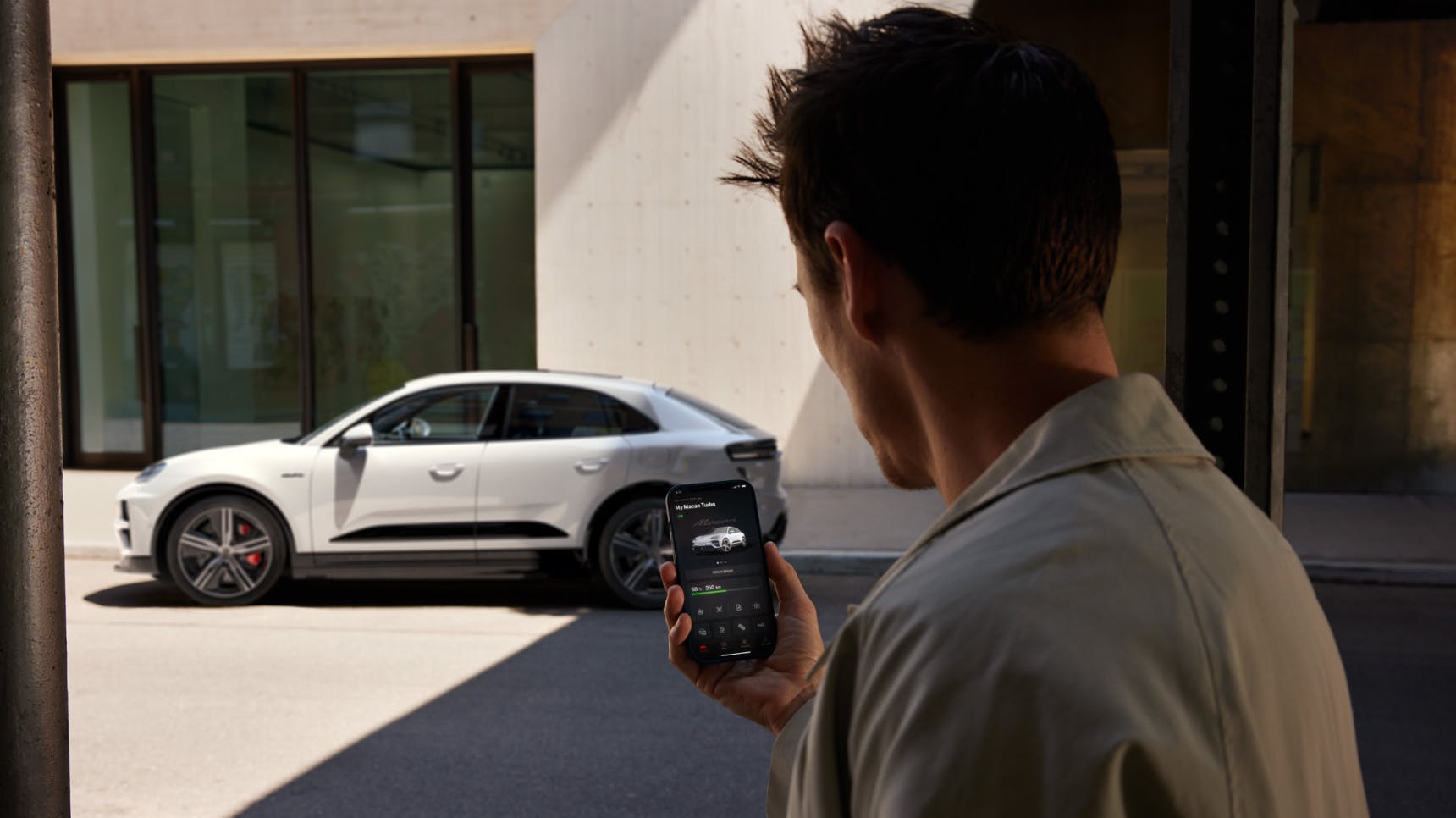 A Porsche Macan Turbo in Ice Gray Metallic is shown parked in front of a building. In the foreground, a man is using his smartphone with the My Porsche app open.