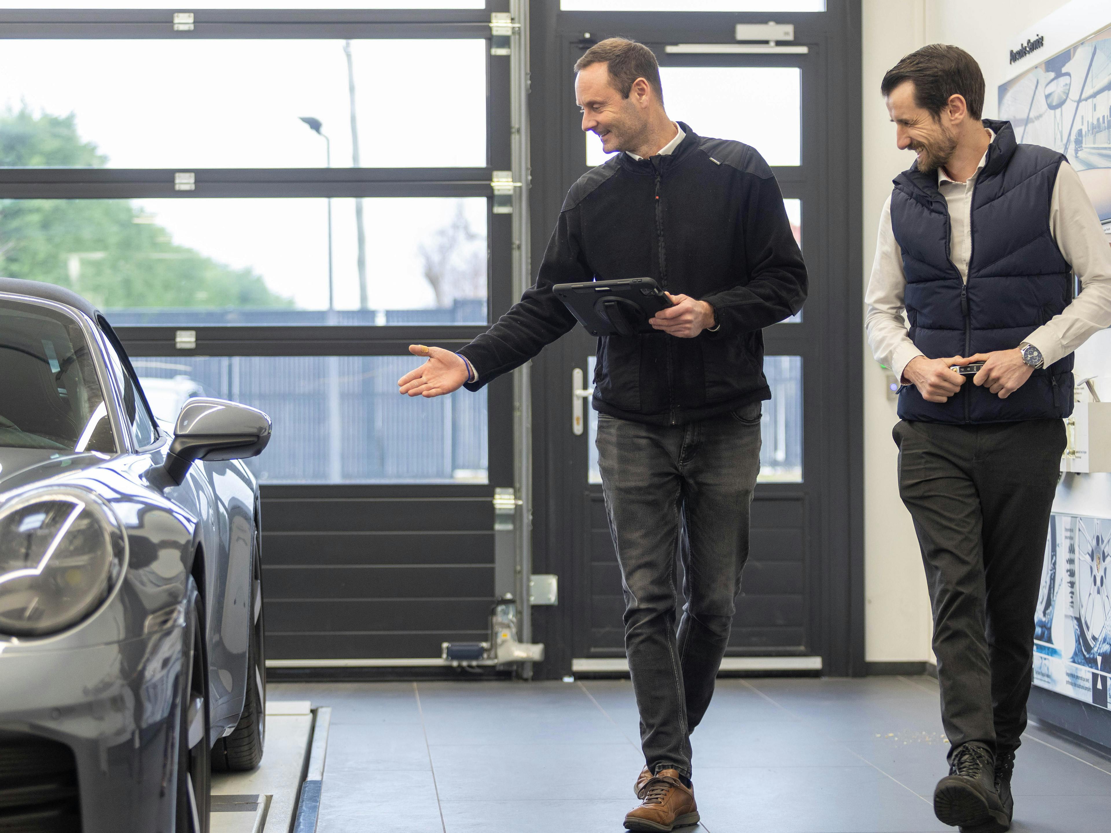 Deux collaborateurs de Porsche passent devant une Porsche grise. L'un d'eux tient une tablette et pointe le véhicule de l'autre main. Son collègue observe également le véhicule.