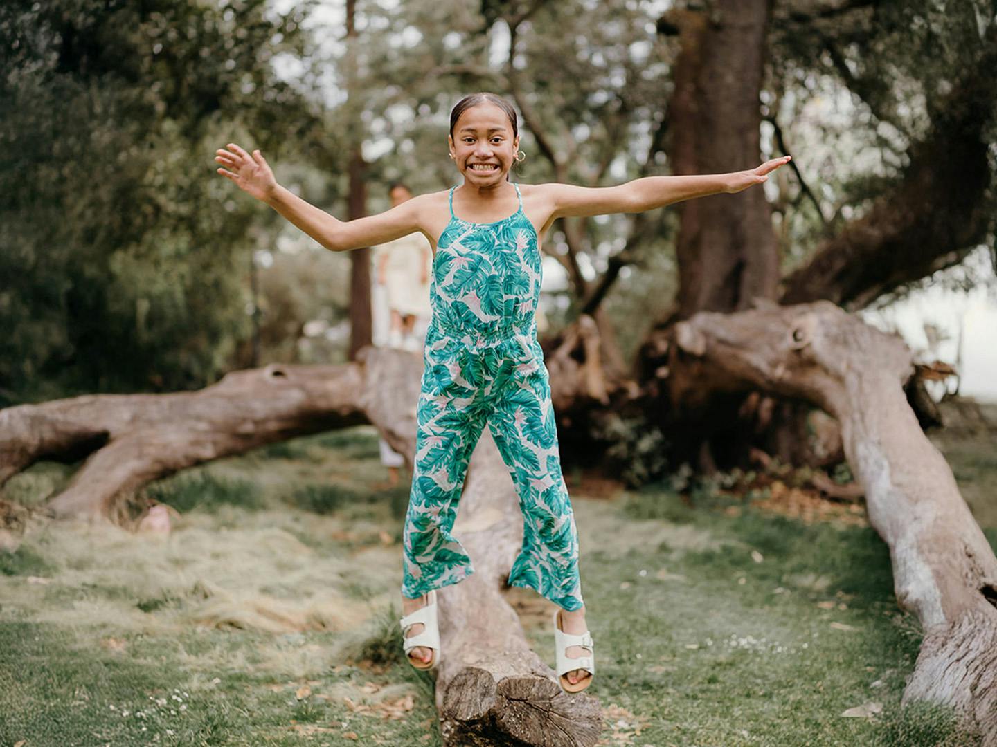 A young girl wearing a green and white patterned sleeveless jumpsuit, stands with arms outstretched in a wooded area.