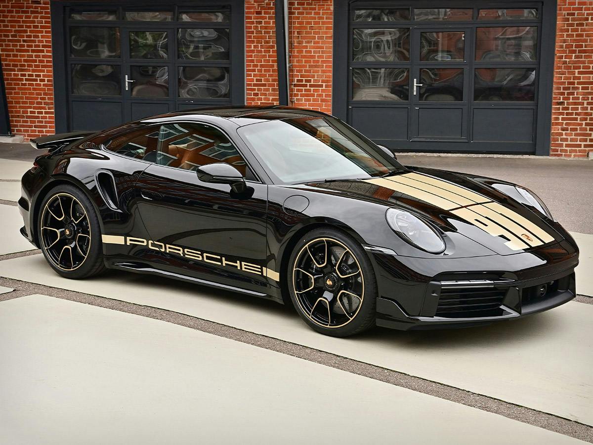Porsche 911 Turbo S Coupe in black, side view, parked on a light-colored surface in front of a brick building with large windows.