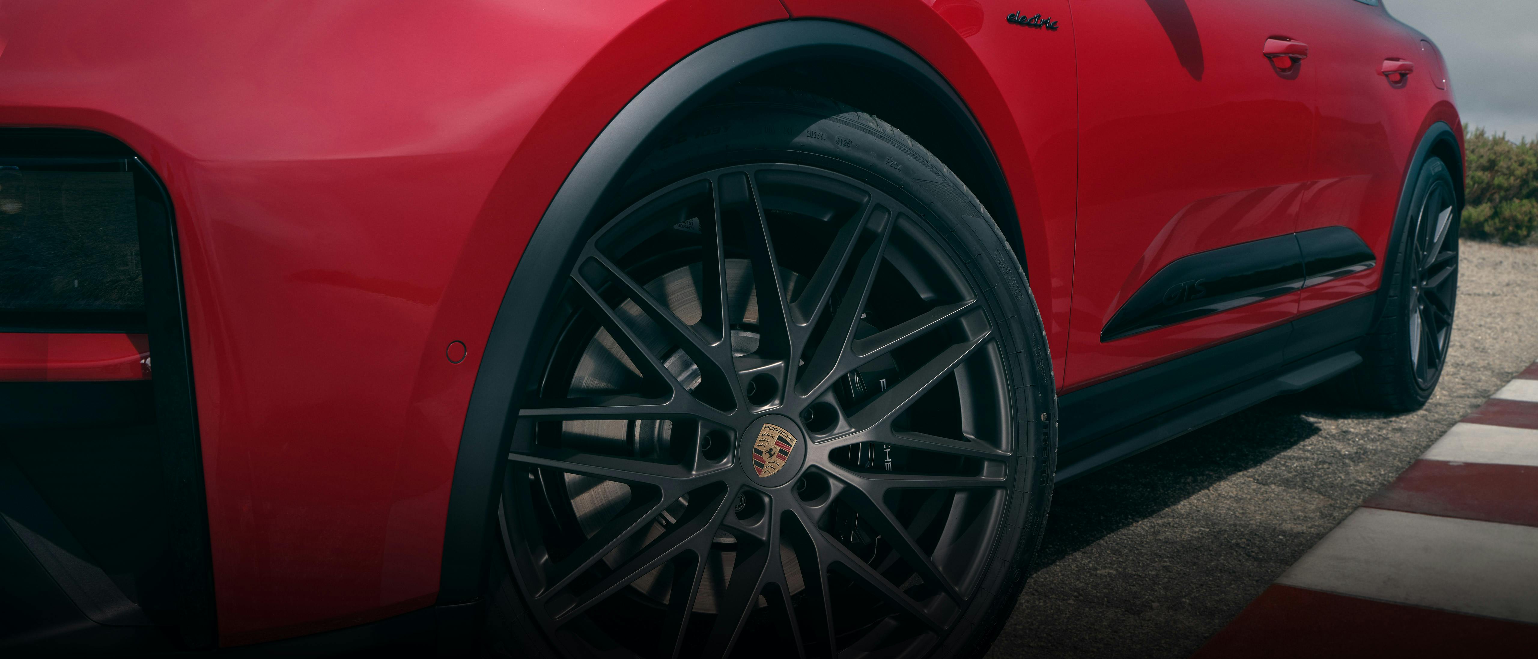 Nahaufnahme eines roten Porsche Macan. Im Vordergrund das vordere Rad mit dunkler Felge und sichtbarem Porsche‑Emblem auf einer Rennstrecke.