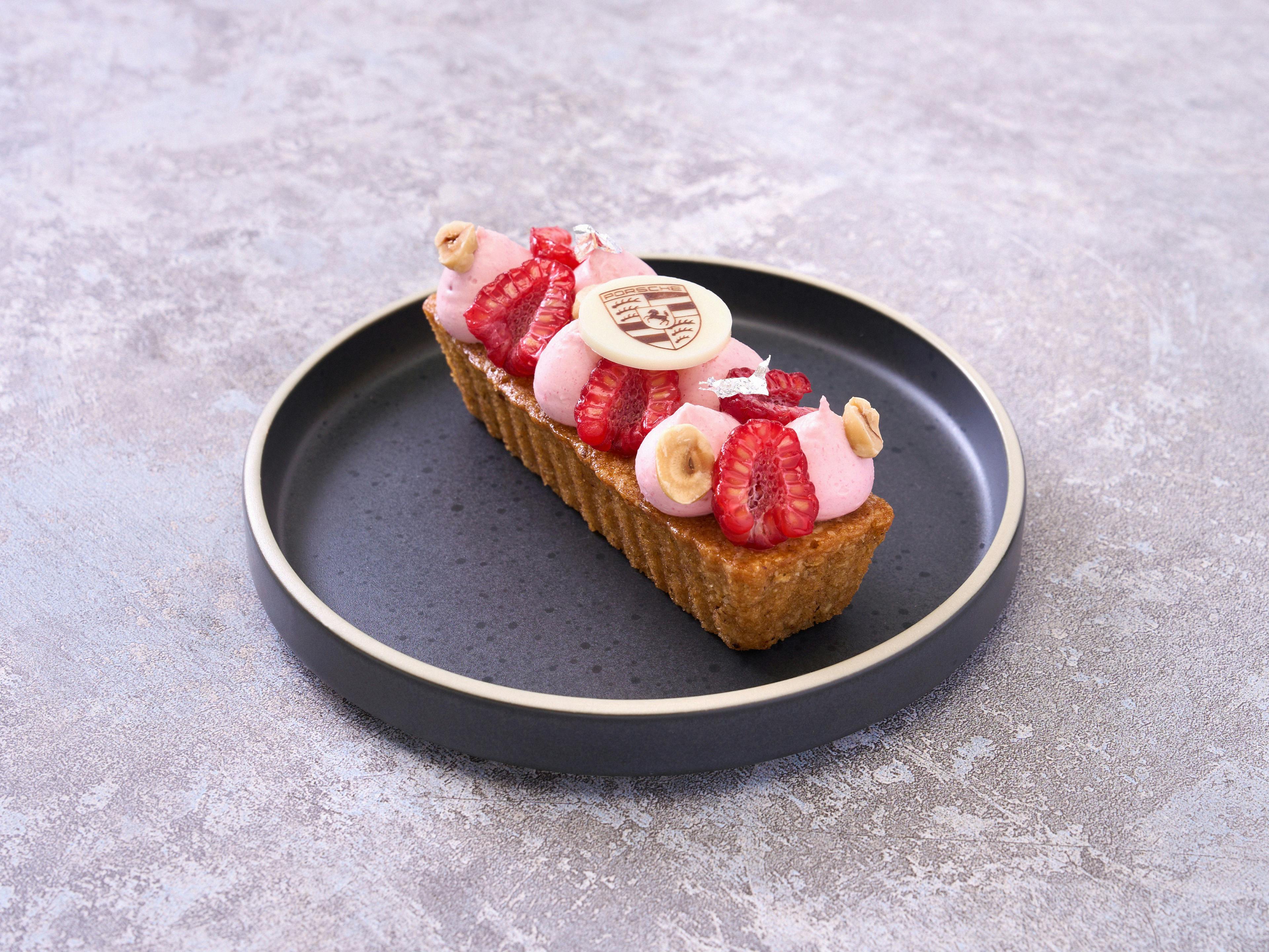 Fruit tart with fresh strawberries, pink cream, hazelnuts and Porsche logo decor on a black plate.