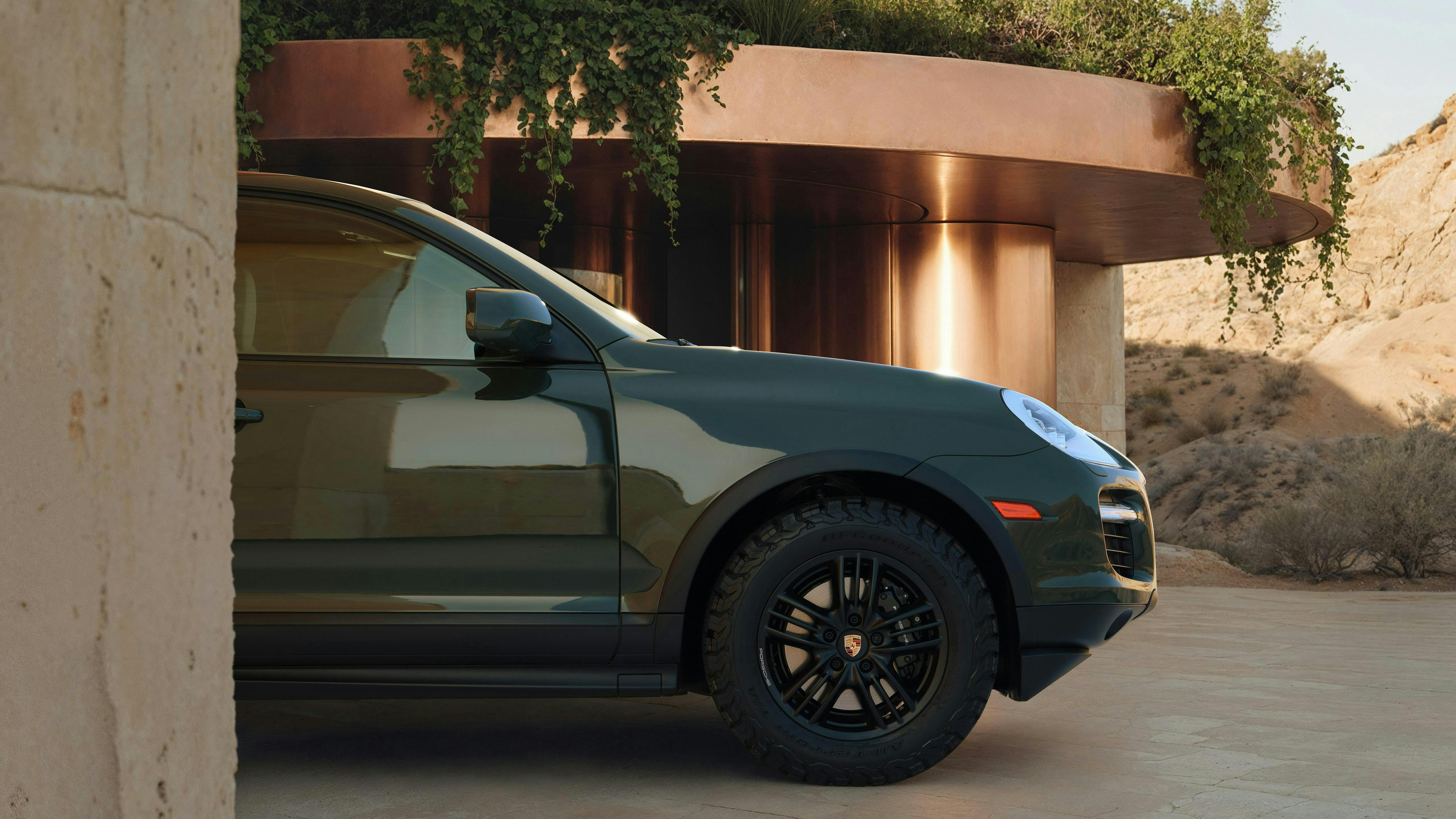 Three-quarter rear view of a Blackolive first-generation Cayenne GTS driving through a sculptural desert-like environment.
