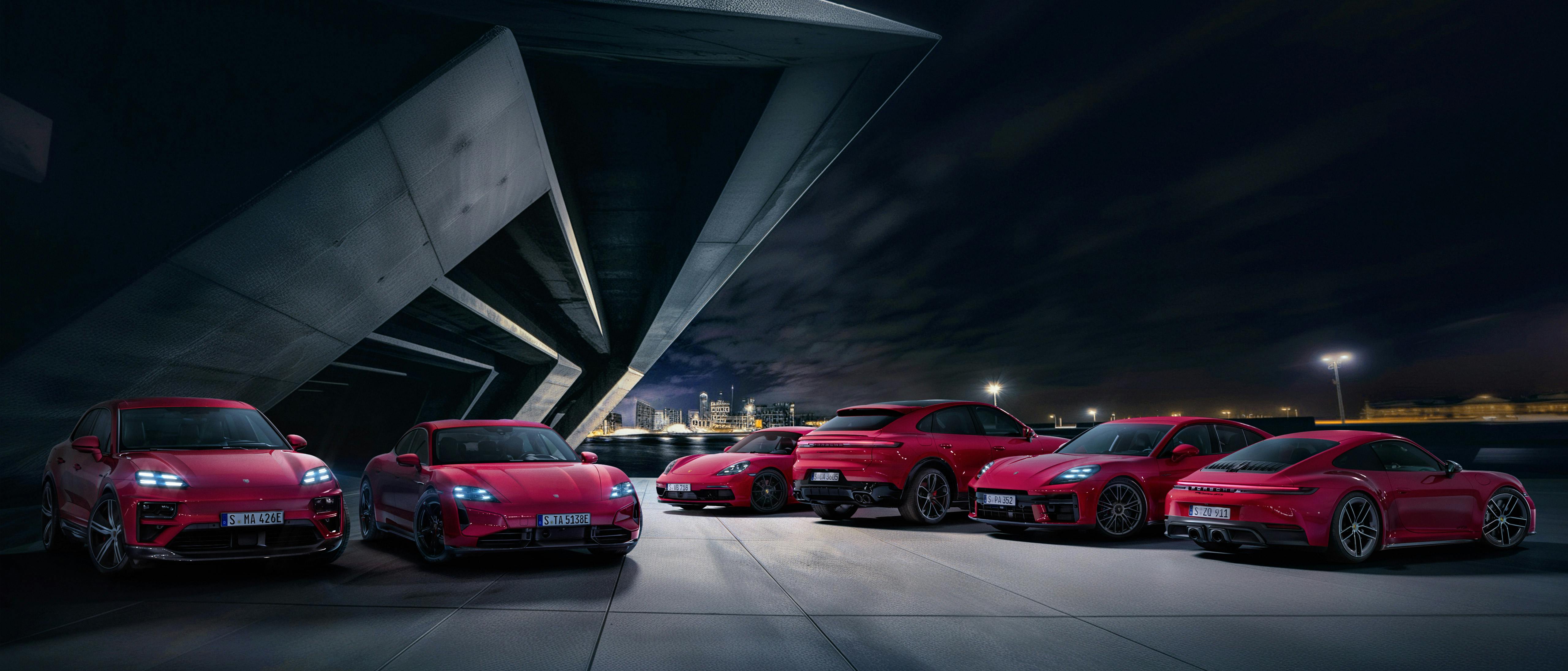 At night, in front of a city full of lights, six different red Porsche models stand under an imposing concrete architecture.