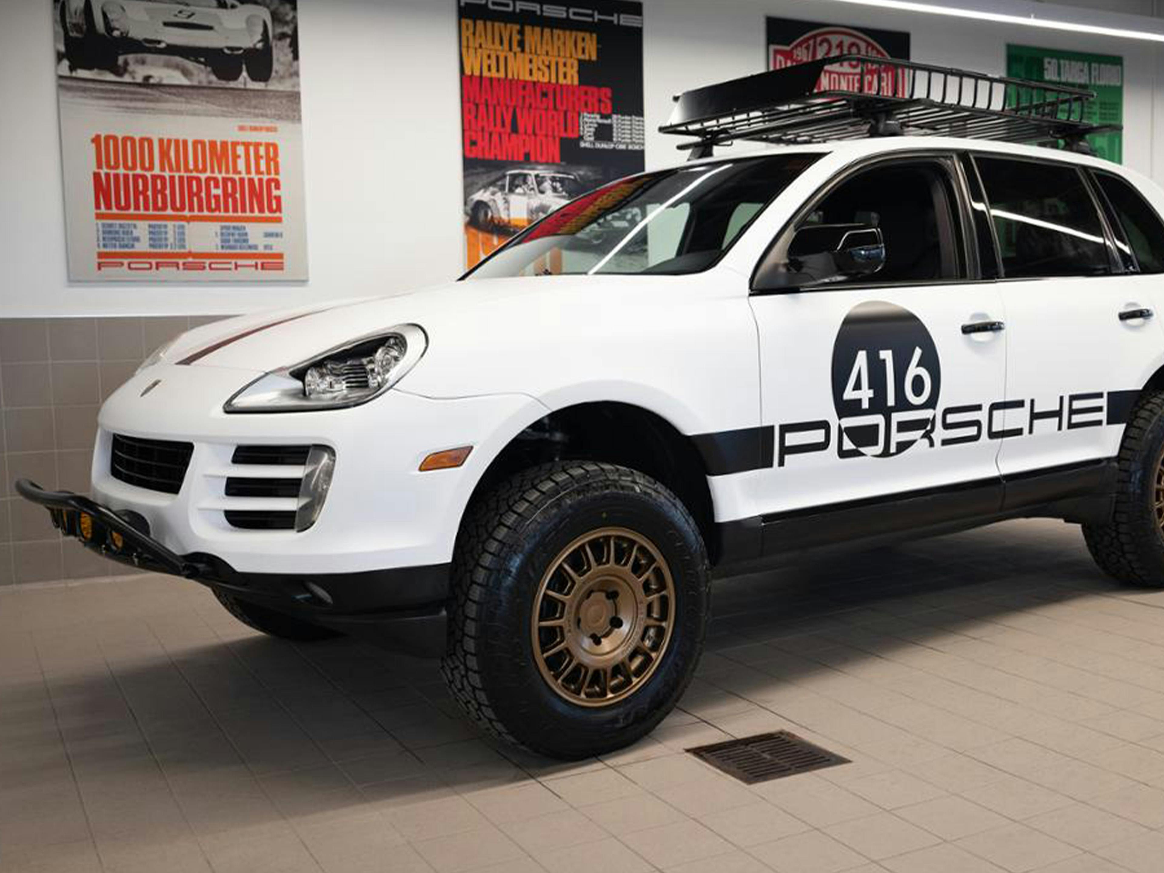 White Porsche SUV with off-road tires and roof rack, labeled “416 Porsche,” parked indoors with racing posters in the background.