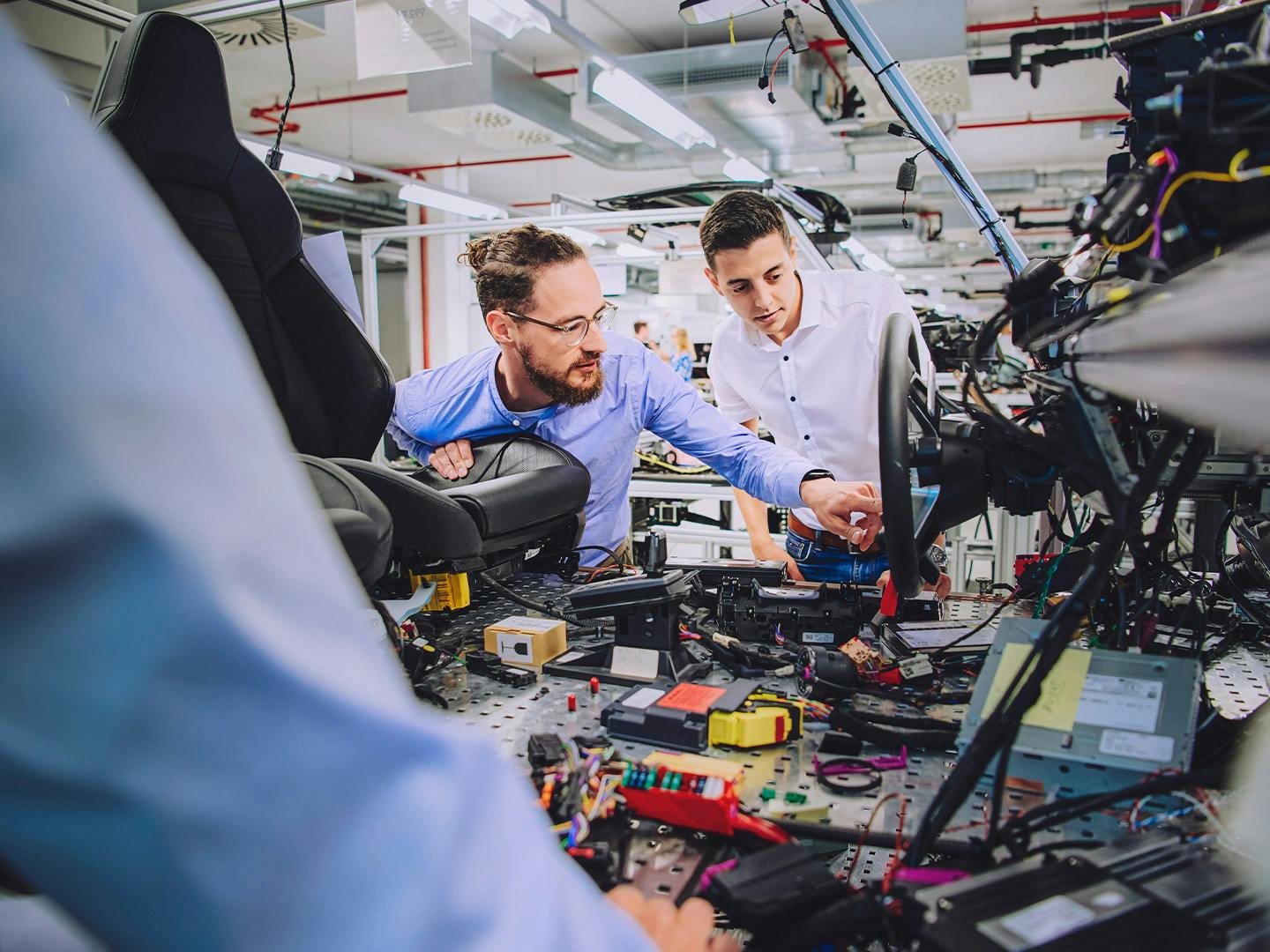 Mehrere Personen arbeiten gemeinsam im Innenraum eines Fahrzeugs in einer modernen Produktionsumgebung, montieren elektronische Bauteile und Kabel und nutzen Werkzeuge sowie Prüfgeräte