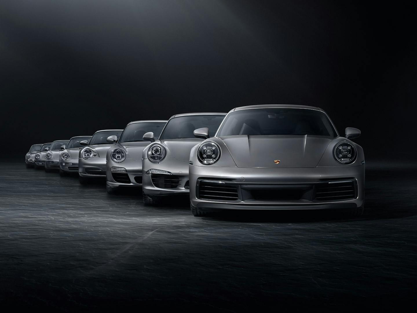 In einem schwarzen Studio stehen verschiedene silberne Porsche 911 Generationsmodelle hintereinander in einer Reihe.