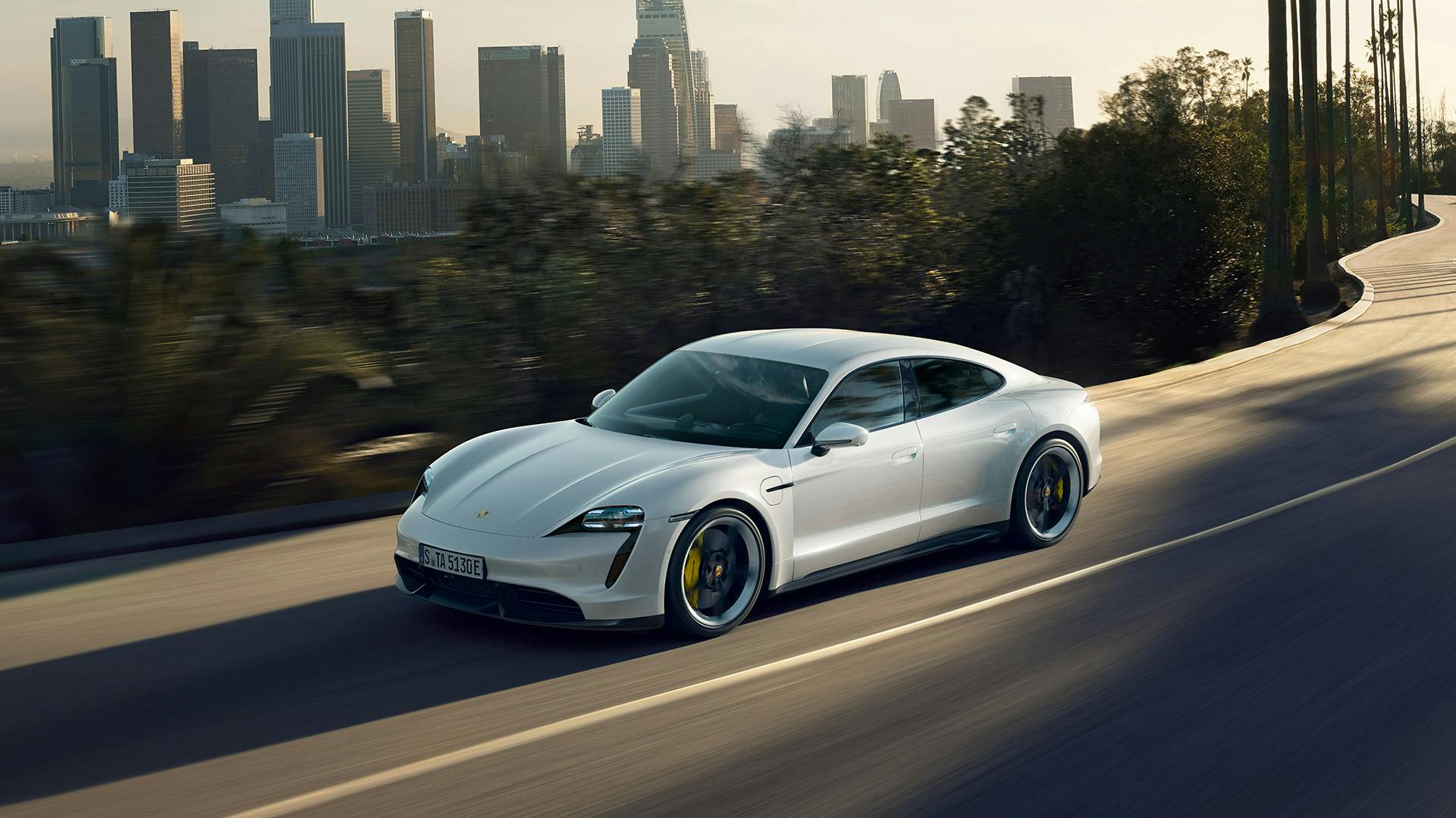 White Porsche Taycan driving on road, with a city panoramic view in the background.