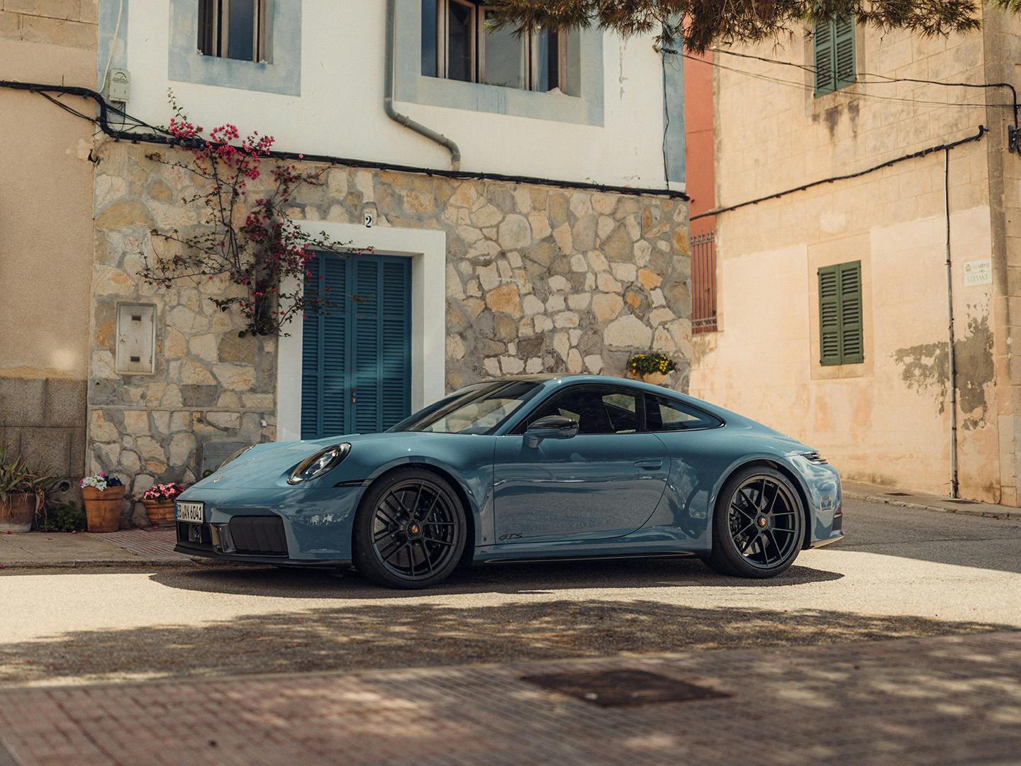 Une Porsche 911 Carrera GTS bleue garée dans une rue calme, devant une maison en pierre rustique avec des volets verts et des pots de fleurs, à l’ombre d’un grand arbre.