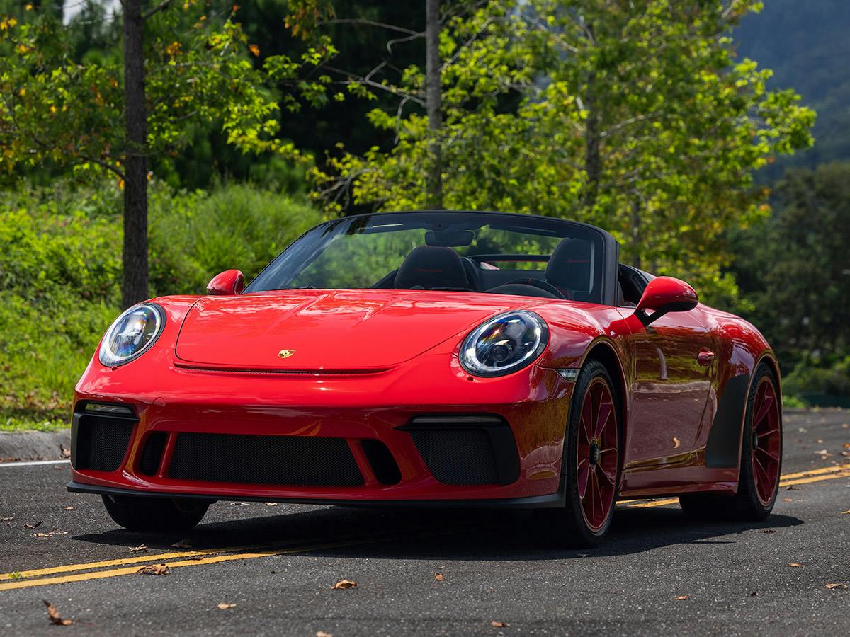 Front three-quarter view of a Porsche 911 Speedster in Paint to Sample colour Guardsred, driving on a tree-lined road with the top down.