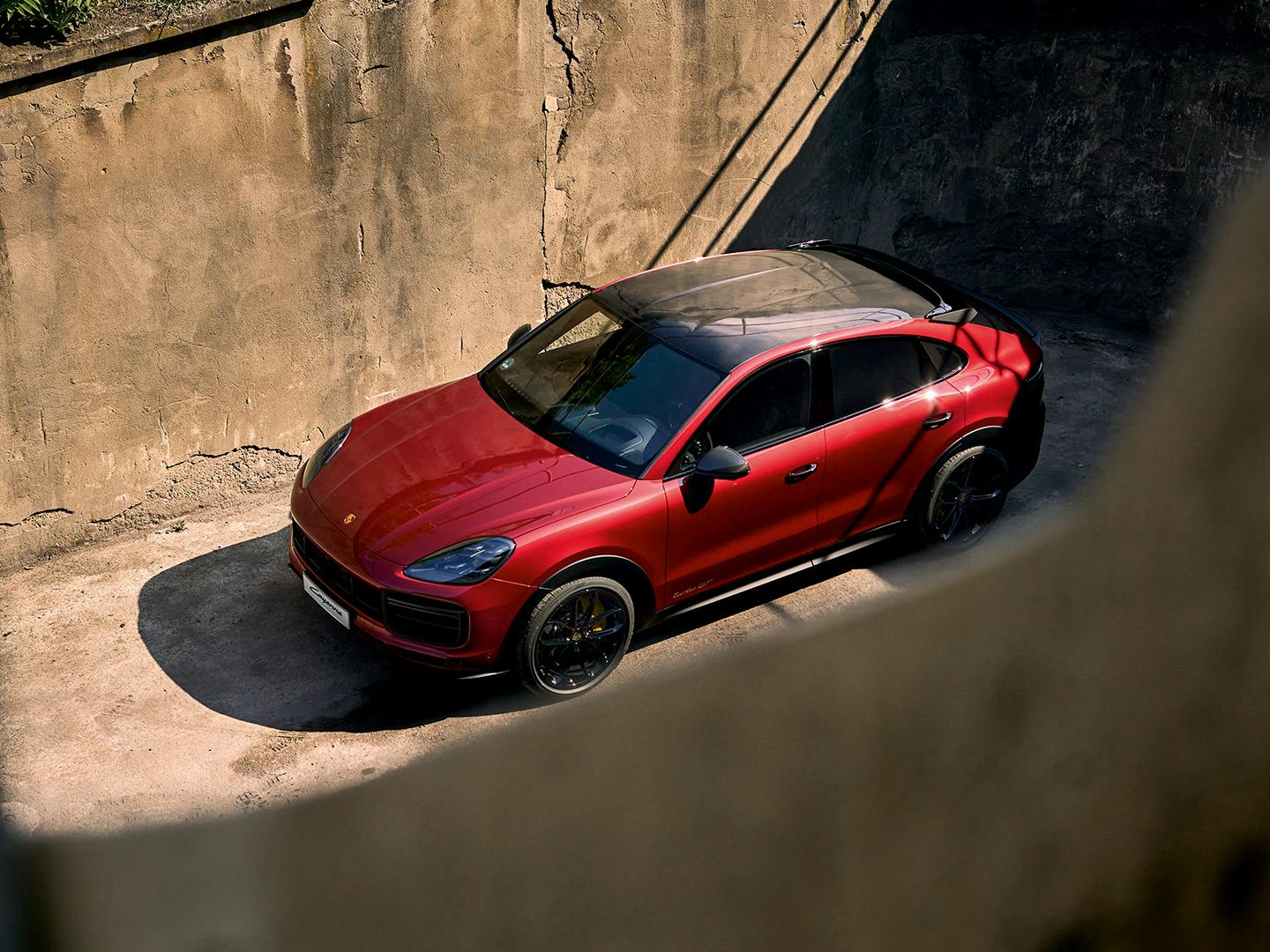 A red Porsche Cayenne Coupe is parked in a sunlit area, surrounded by tall concrete walls. The car features a sleek, sporty design with black accents, tinted windows, and a panoramic glass roof.