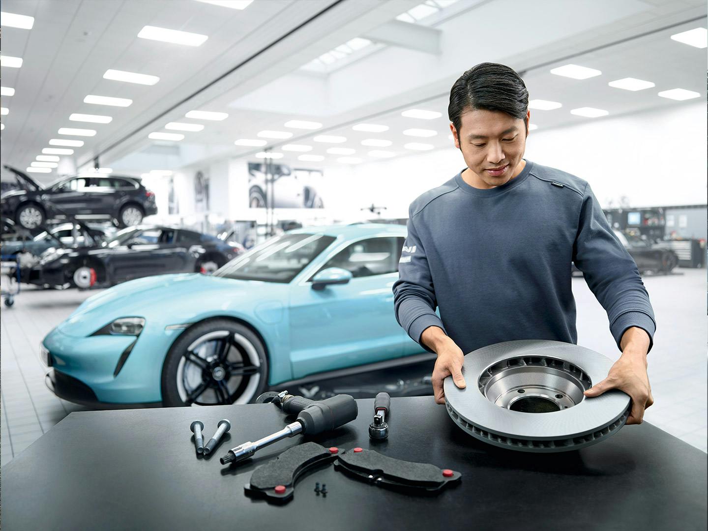 A Porsche technician inspecting a brake disc in a modern workshop, with a Turquoise Porsche Taycan in the background