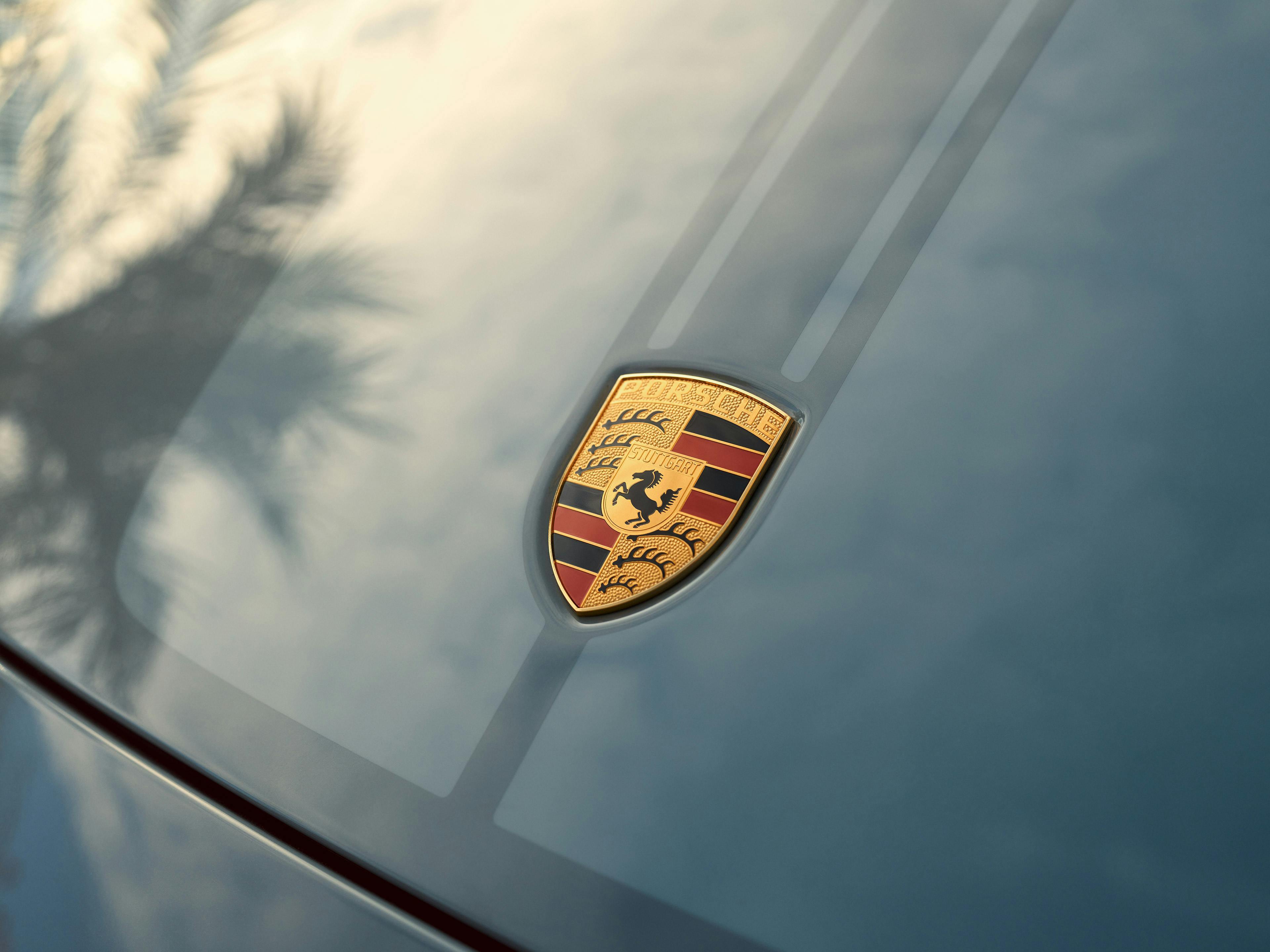 Ein Porsche-Emblem geprägt auf einer blauen Motorhaube, überschattet von den Silhouetten der Bäume.