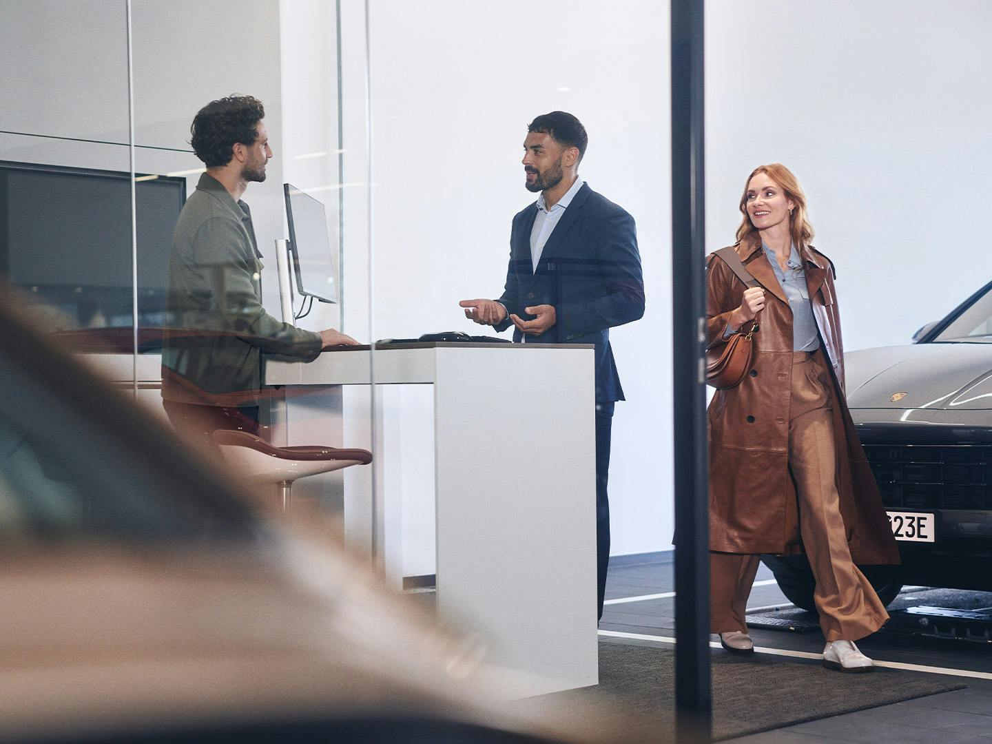 At the reception of a Porsche service centre, a customer is talking to a service advisor. A woman walks past in the background.