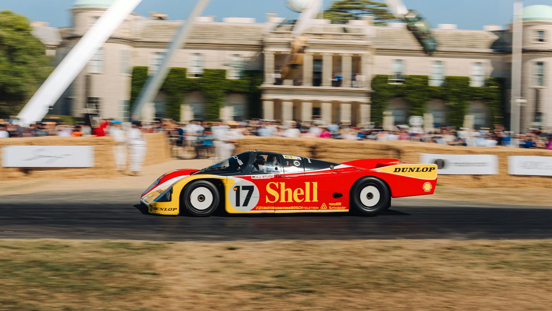 Liveried Porsche race car climbing the hill in front of Goodwood House.
