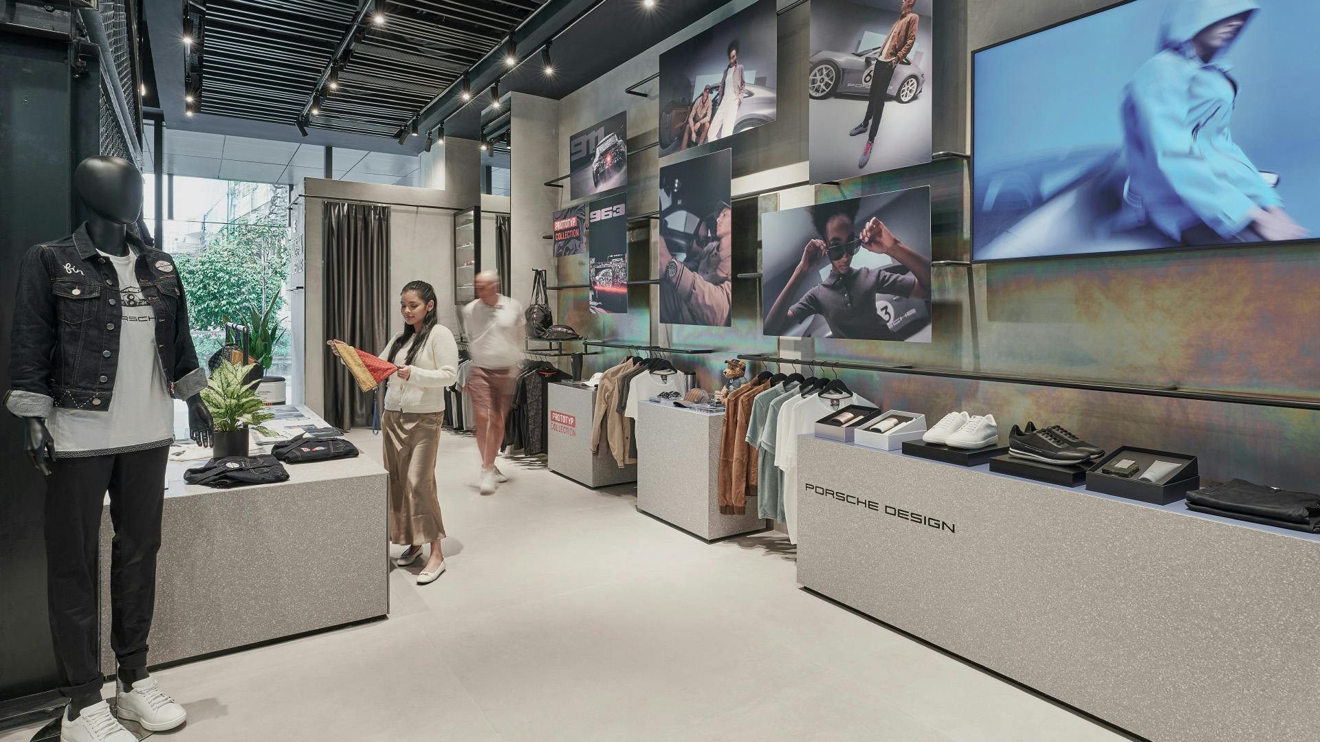 Modern Porsche lifestyle store with fashion displays, digital screens, and customers browsing clothing and accessories.