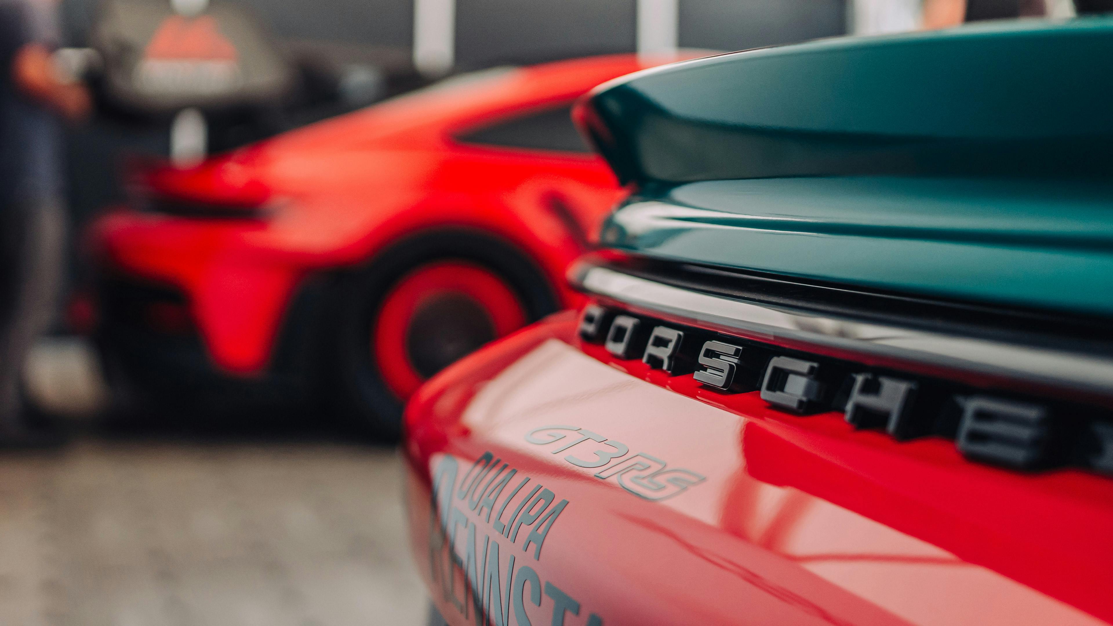 A close-up of the Porsche lettering on a teal Porsche, with a red car blurred in the background.