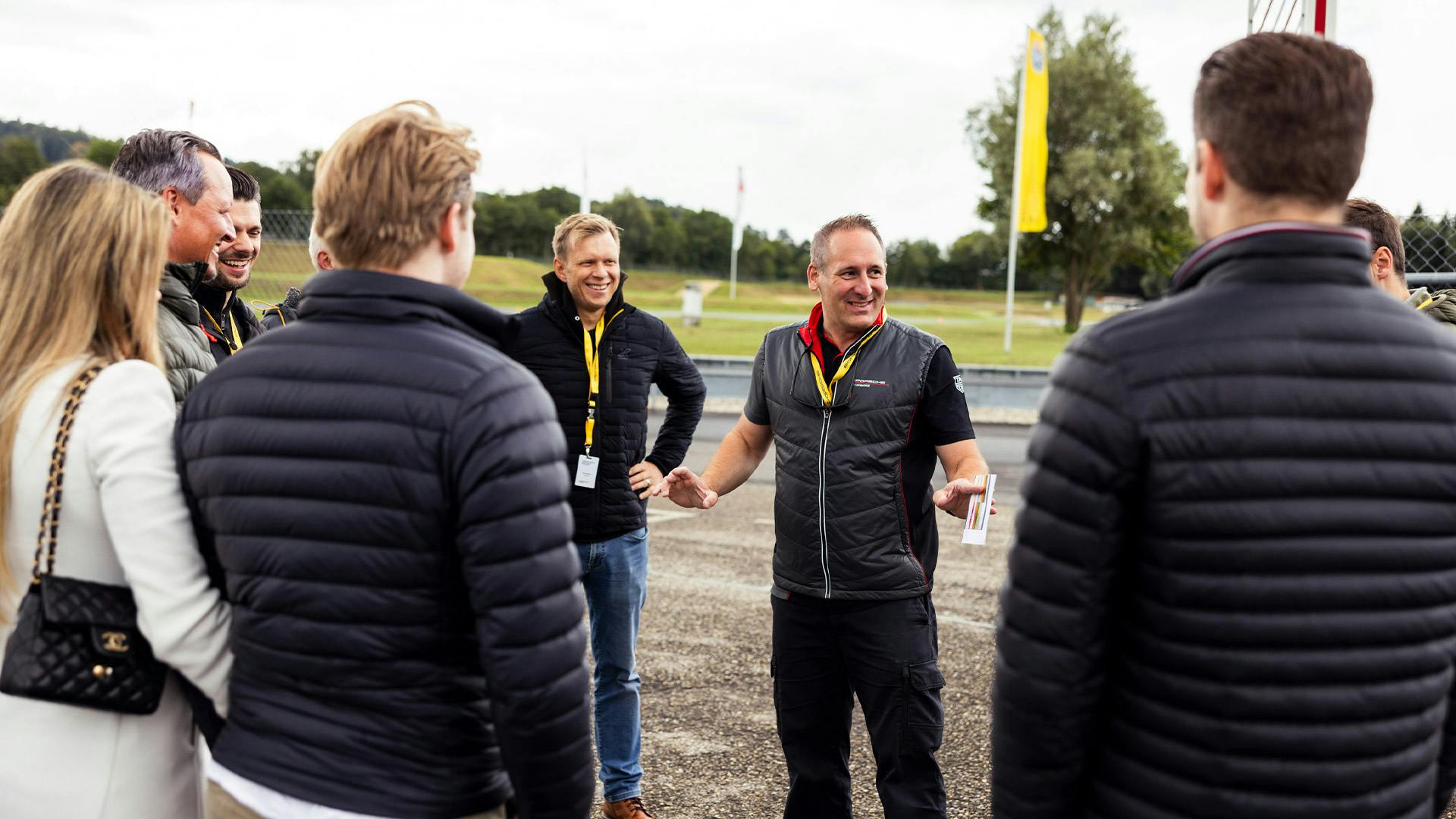 Ein Mann in einer grauen Porsche Experience Weste steht am Rand der Rennstrecke, umgeben von einer Gruppe gut gelaunter Besucher.