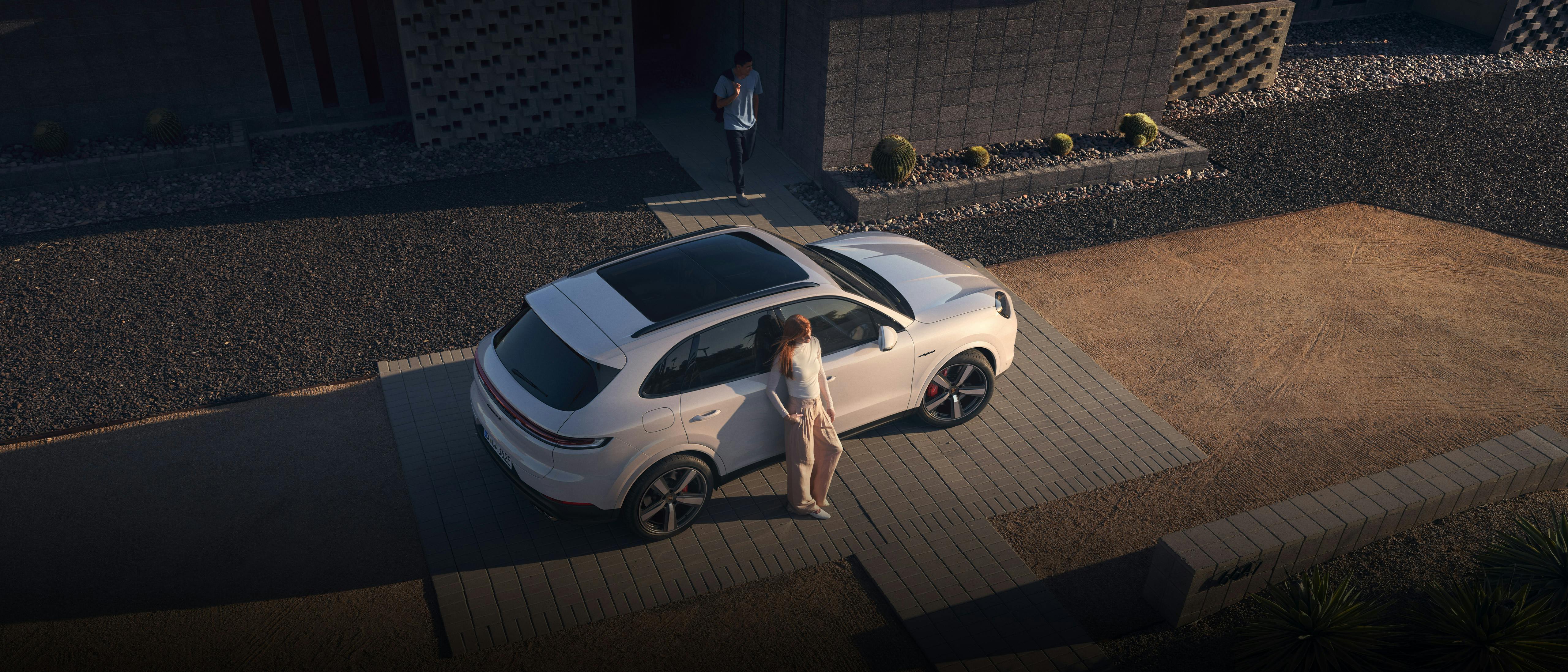 Luftaufnahme eines modernen Wohnhauses. Ein weißer Porsche Cayenne steht in der gepflasterten Einfahrt, eine Person neben der geöffneten Fahrertür.