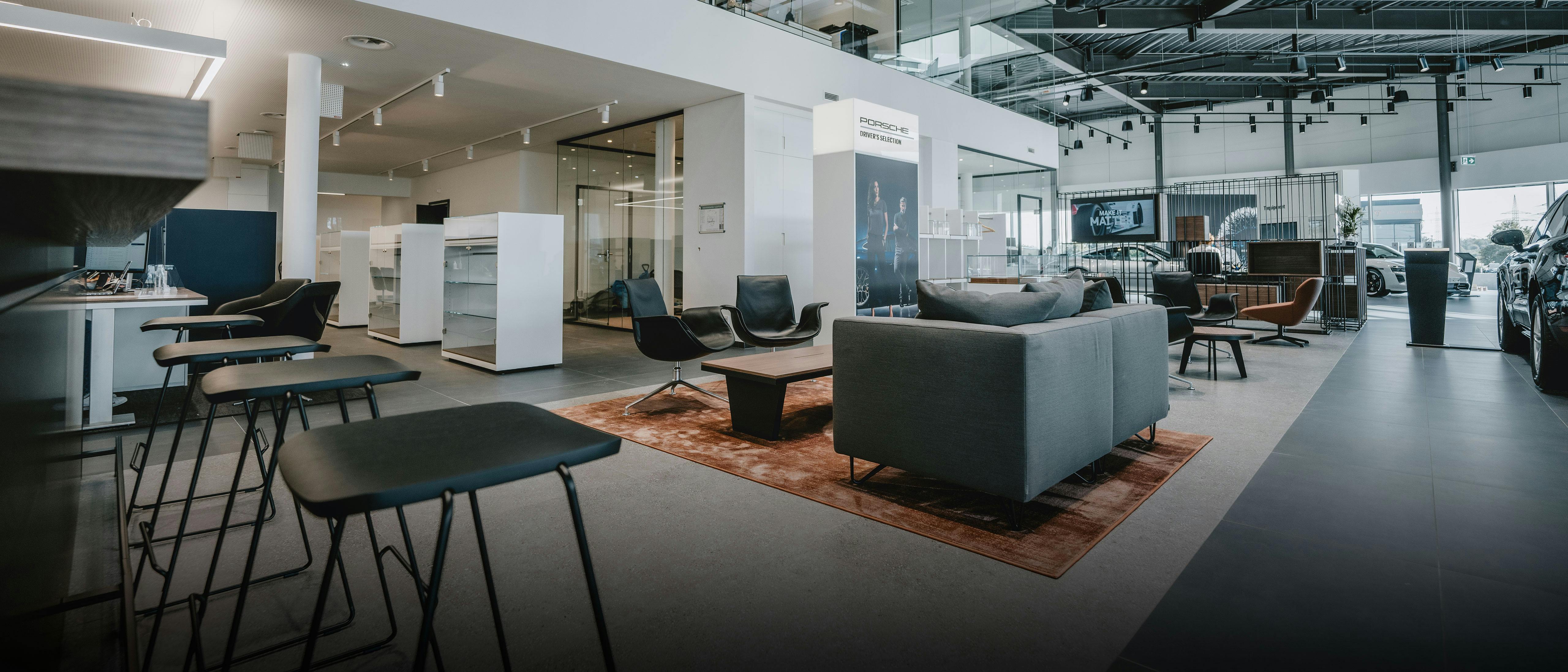 Vue du showroom moderne du Porsche Zenter Roost, avec différentes options de sièges pour les clients, dans des tons de blanc, gris et brun.