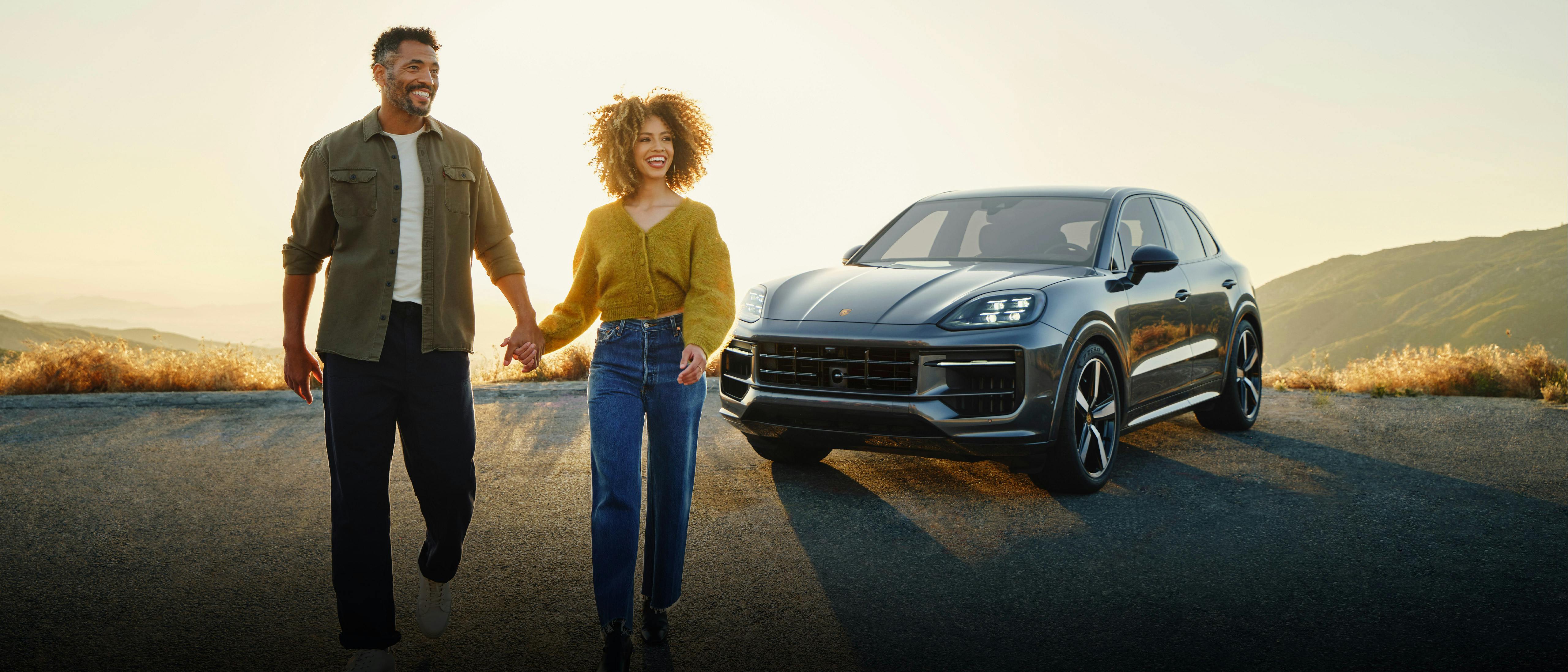 A smiling couple hold hands in front of a Porsche Cayenne at sunset in the mountains.