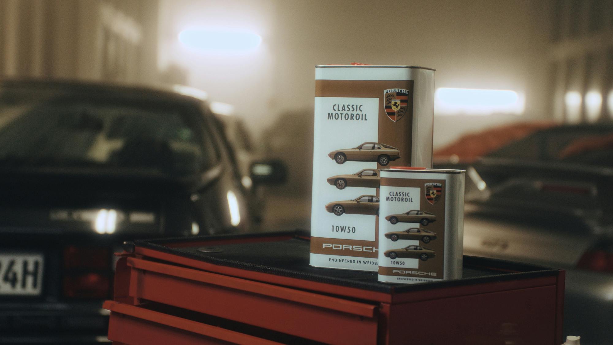 Two rectangular cans of Porsche Classic Motoroil 10W-50 sitting on top of a red metal tool chest in a dimly lit garage. The cans feature images of vintage sports cars and the Porsche crest. The background is showing the silhouette of a classic car parked in the garage.