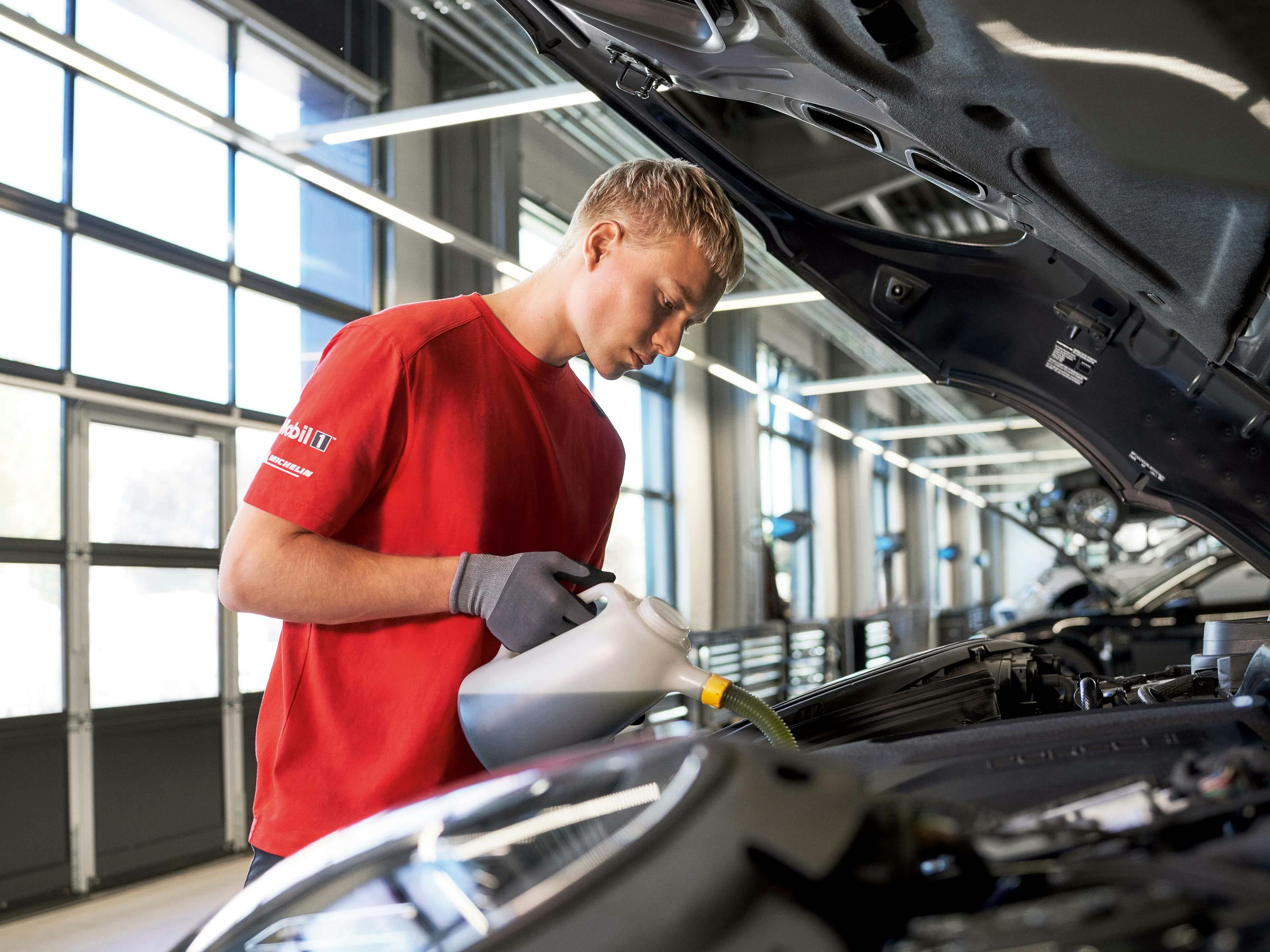 Een man in een rode werkplaats Porsche polo giet vloeistof uit een kan in de motor van een auto. Hij draagt een grijze handschoen en kijkt geconcentreerd naar wat hij doet. De motorkap van de auto is open.