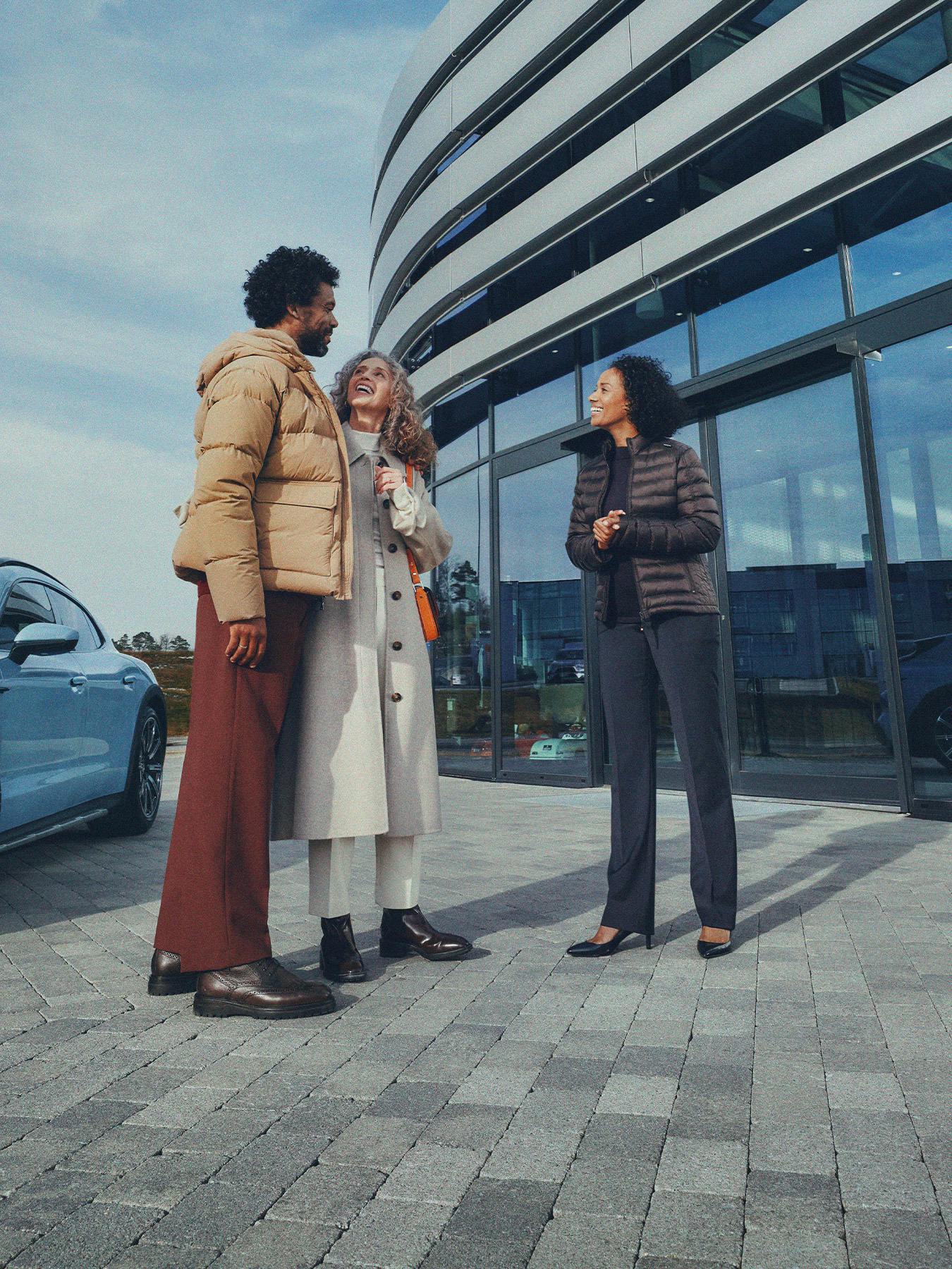 Three people in conversation beside a blue Porsche outside the curved glass façade of a Porsche Centre.​