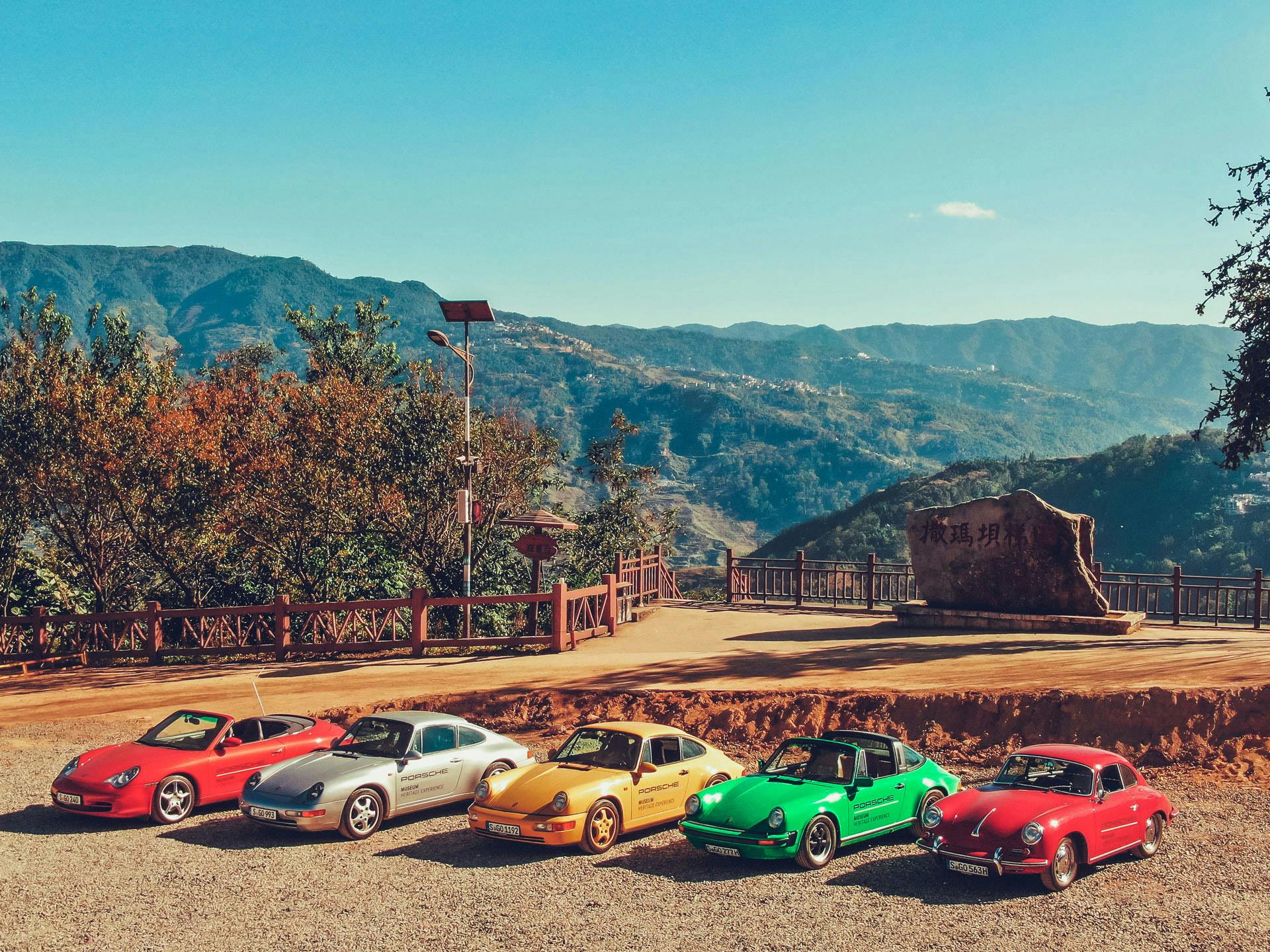 Several different coloured Porsche vehicles on a gravel surface in front of a hilly landscape