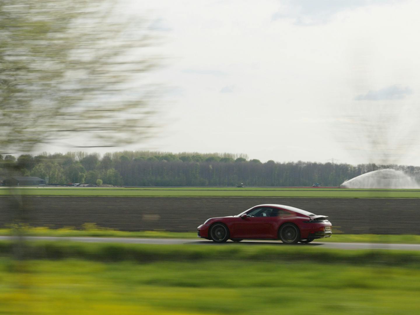 Dynamisch zijaanzicht met bewegingsonscherpte van een rode Porsche 911 die langs een bloembollenveld en een waterberegeningsinstallatie rijdt. Het zijprofiel van de sportauto komt duidelijk naar voren in dit actie-shot in een typisch Nederlands landschap.
