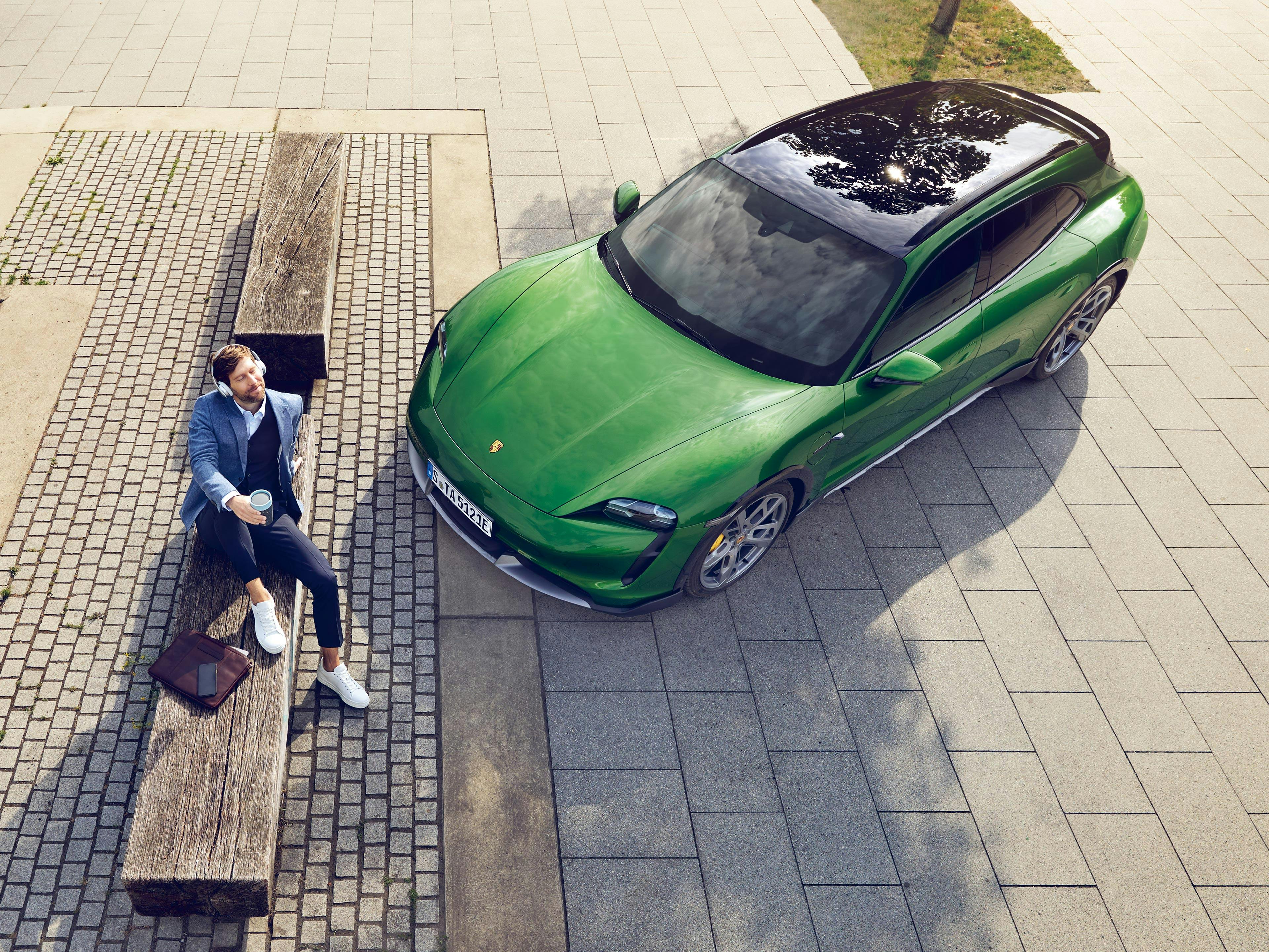 Ein Mann mit Coffee-to-Go-Becher sitzt auf einer Bank neben einem Brunnen, daneben parkt ein grüner Porsche.