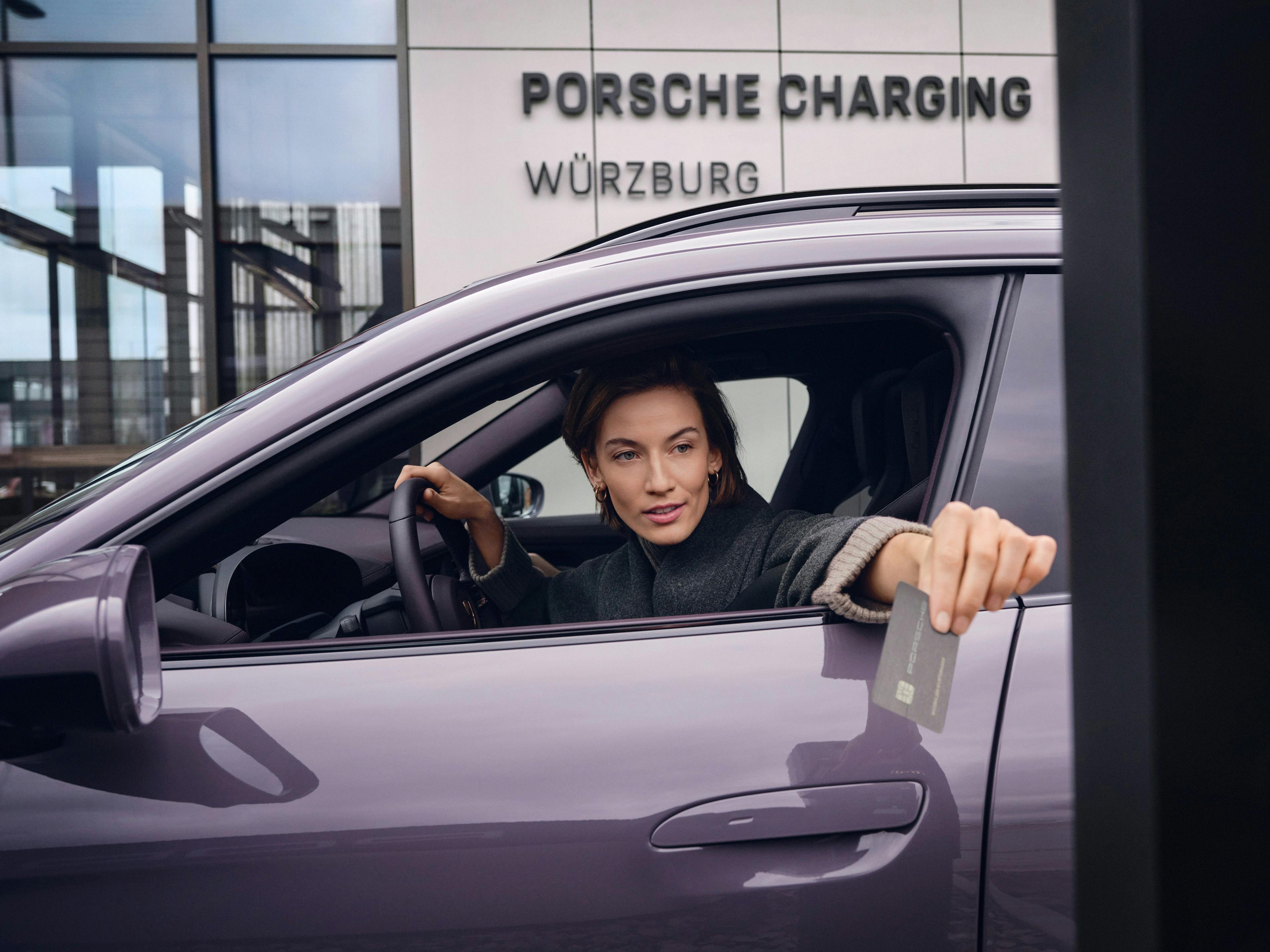 Eine Frau lehnt sich aus ihrem violettfarbenem Sportwagen und hält eine graue Karte in der Hand. Im Hintergrund ist ein Gebäude zu sehen mit dem Schriftzug "Porsche Charging Würzburg".
