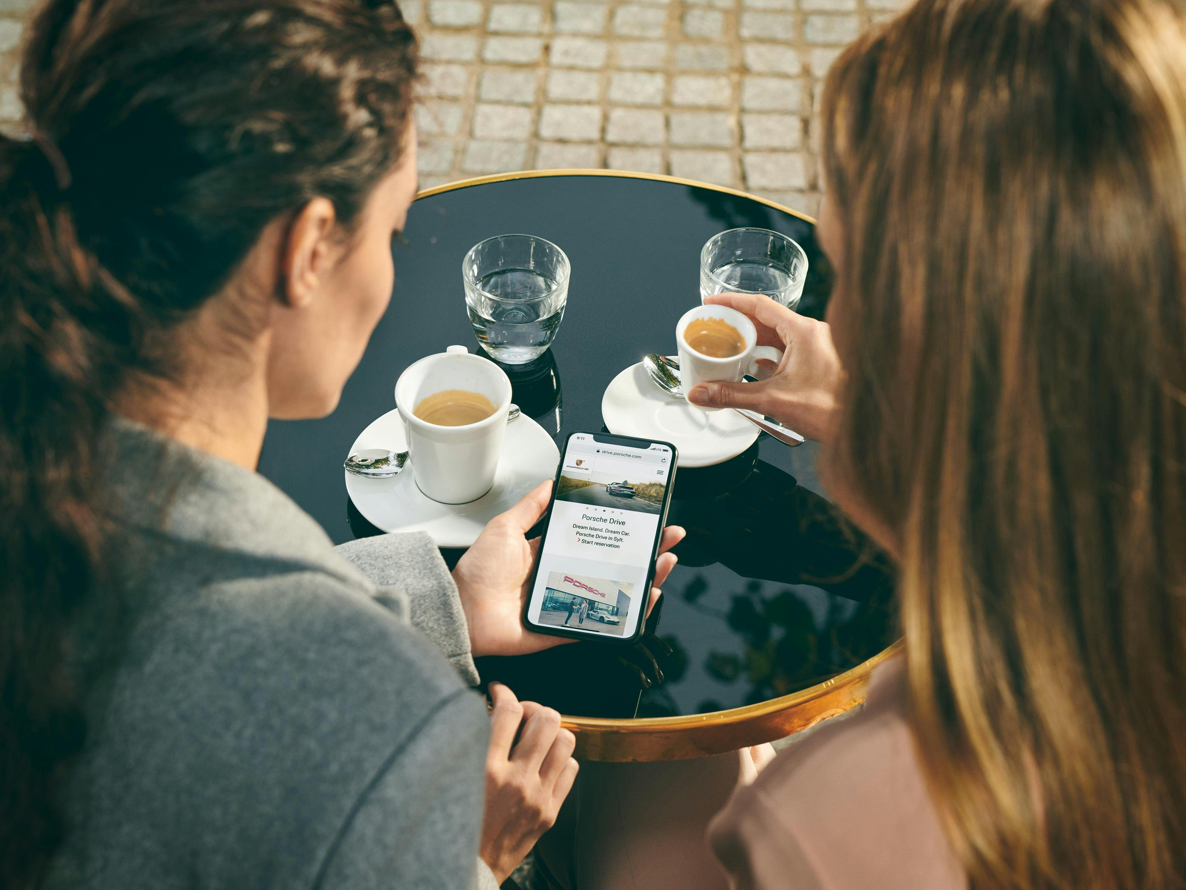 2 人の女性がコーヒーを飲みながら小さな丸いテーブルに座り、一緒に携帯電話を見ている。