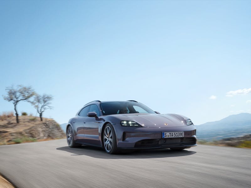 A Porsche Taycan driving on a country road. Bare trees and a clear sky can be seen in the background.