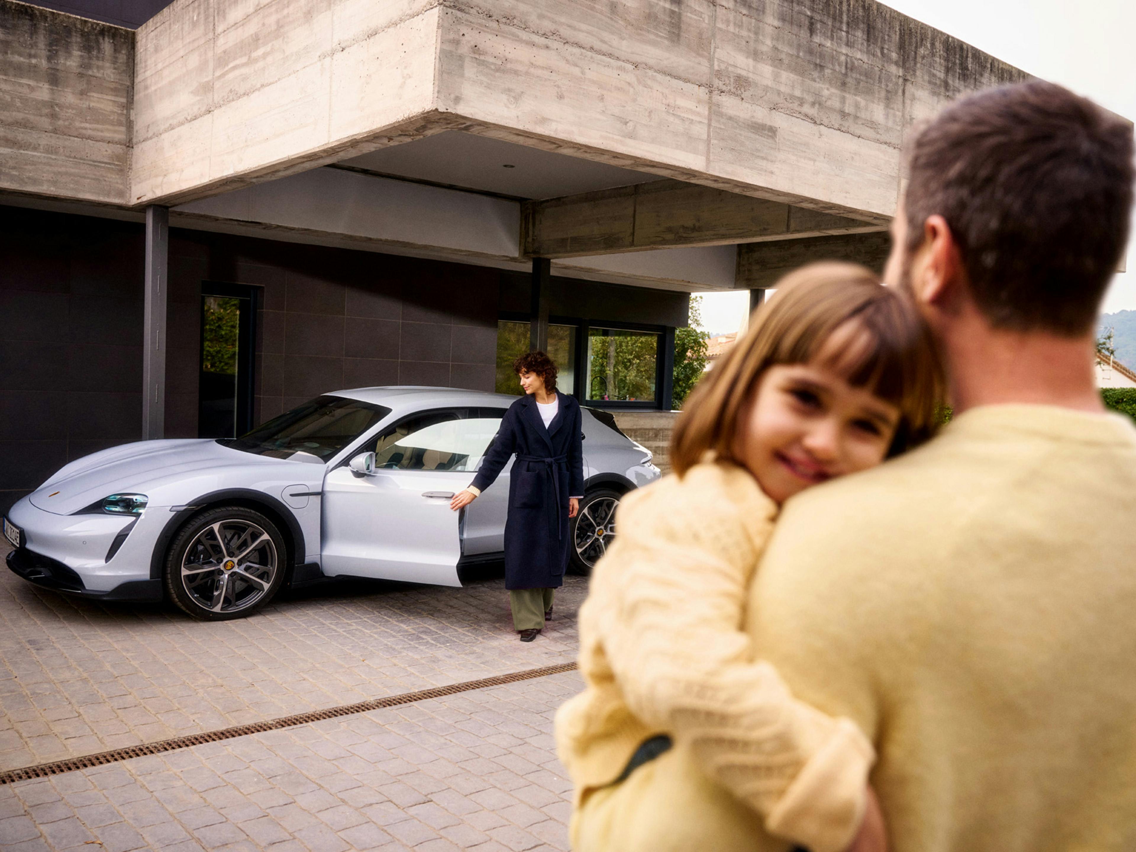 Ein Vater hält seine Tochter und beobachtet seine Frau, wie sie in den Porsche Taycan steigt.