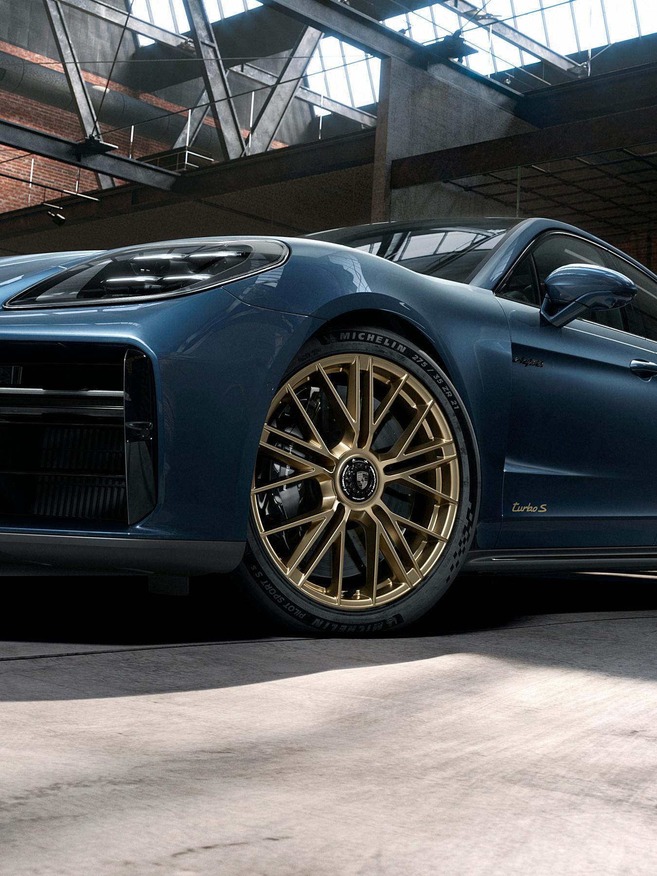 A low-angle and close-up shot of a blue Porsche Panamera Turbo S parked in a modern industrial garage. The shot emphasises the intricate gold wheels.