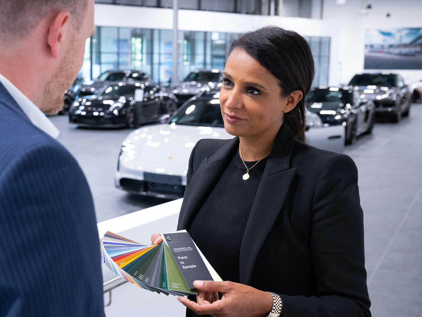 Porsche-Beraterin zeigt Lackfarbfächer einem Kunden im Showroom mit schwarzen Modellen im Hintergrund.