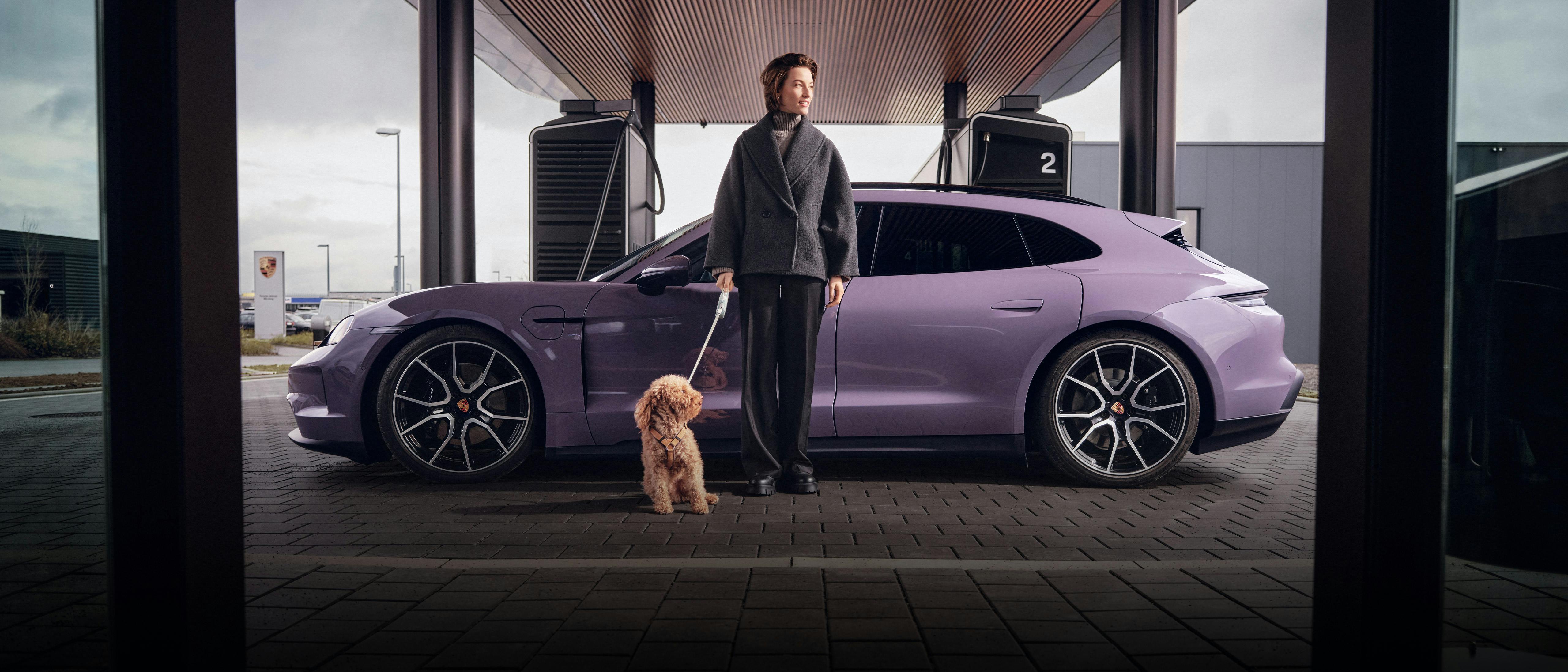 A woman in a Grey coat standing next to a Porsche Taycan at a charging station. She is holding a small dog on a leash.